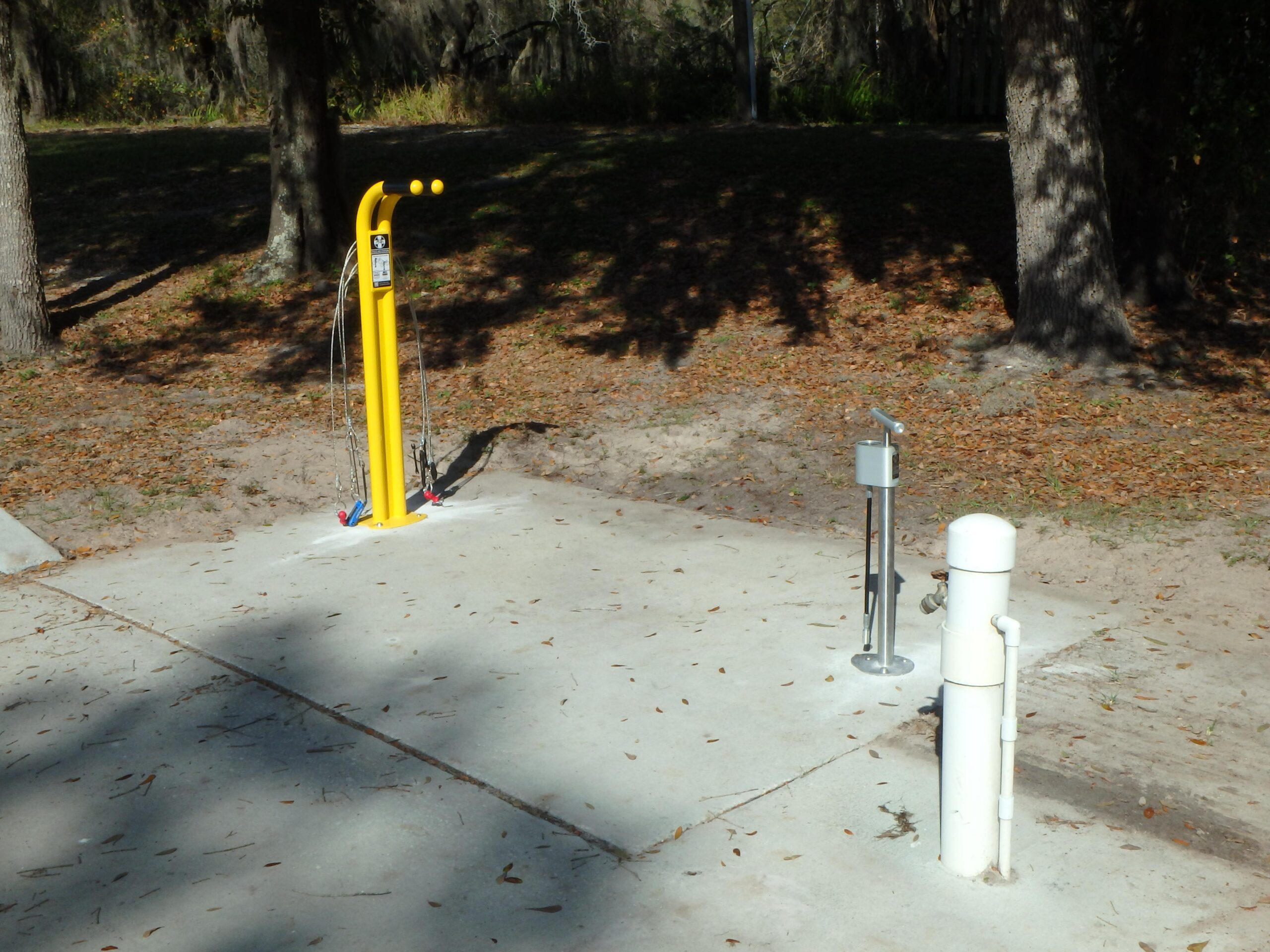 Yellow outdoor fitness equipment with two handles and attached cables on a concrete pad, surrounded by shaded trees and fallen leaves. Two visible water pump fixtures are located nearby. Alafia River State Park mountain bike trail.