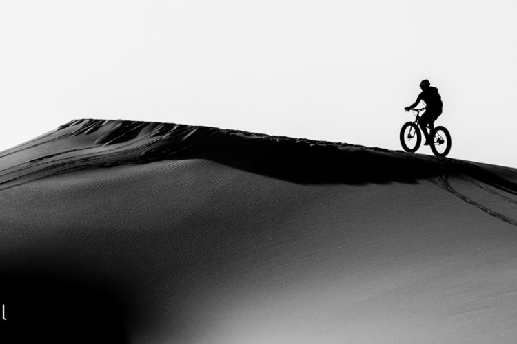 A silhouetted mountain biker riding along the crest of a large sand dune, captured in black and white. The rider is positioned against a bright sky, emphasizing the contrast of the smooth, flowing lines of the dune.