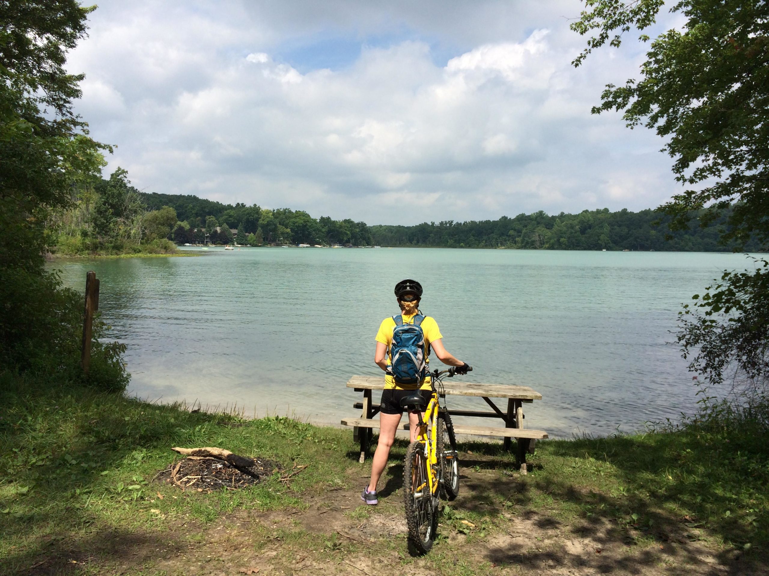 A person in a yellow shirt and black bike helmet stands beside a bicycle, gazing out at a tranquil lake surrounded by greenery. A picnic table is visible nearby, and the sky is partly cloudy, enhancing the serene outdoor atmosphere. Potawatomi trail mountain bike trail.