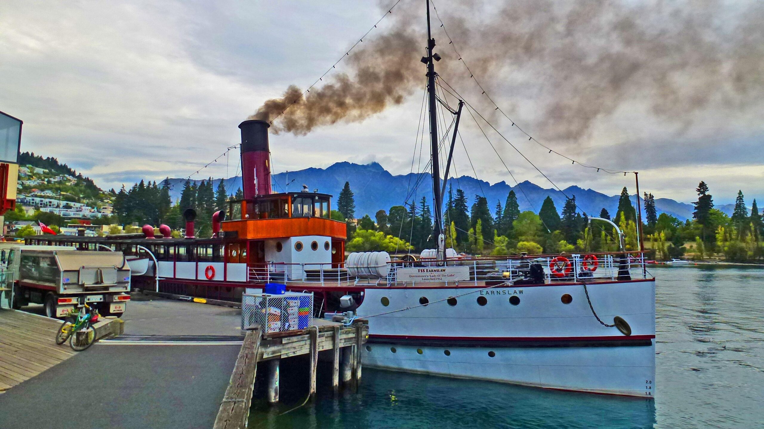A vintage steamship, the TSS Earnslaw, docked at a waterfront with a backdrop of mountains and trees, emitting black smoke from its chimney. A bicycle leans against a wooden railing on the pier. The scene is set under a cloudy sky, depicting a serene lakeside atmosphere. Mavora Lakes mountain bike trail.