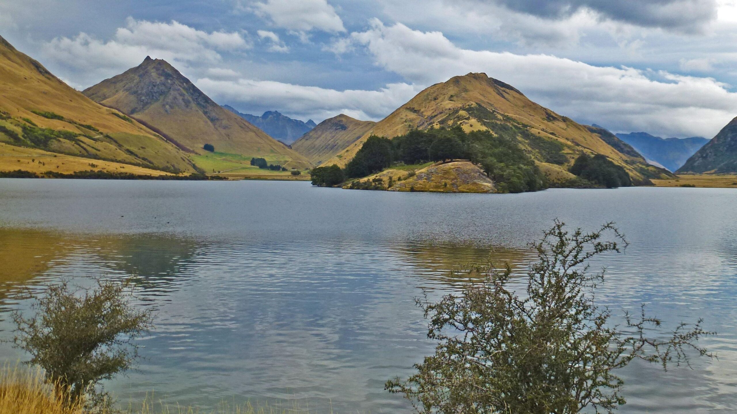 A serene landscape featuring a calm lake reflecting surrounding mountains and hills, with a mix of grassy slopes and patches of trees. The sky is overcast with soft clouds, adding to the tranquil atmosphere of the scene. Moonlight Circuit mountain bike trail.