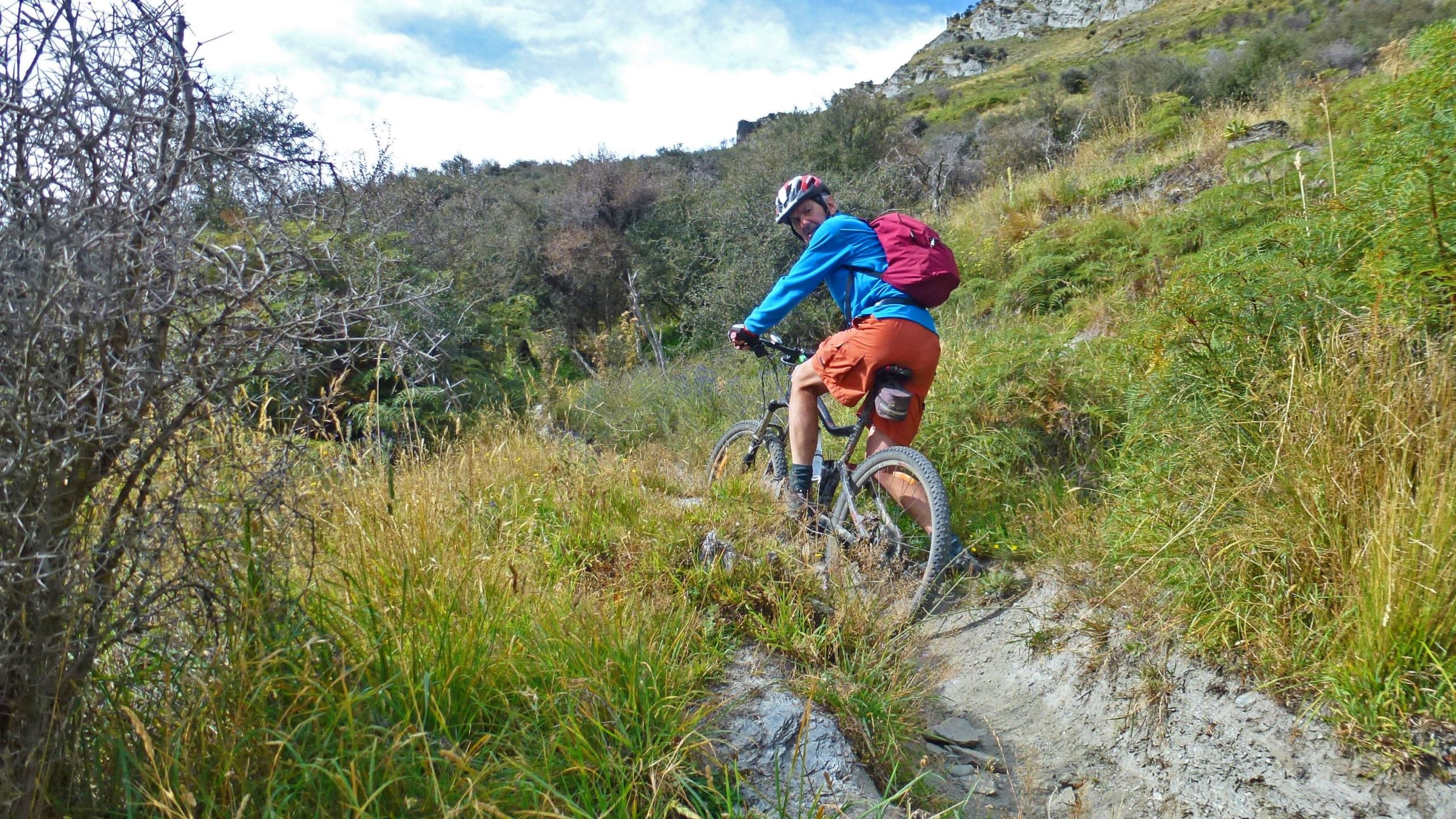 A person riding a mountain bike on a dirt trail surrounded by tall grass and greenery. They are wearing a blue long-sleeve shirt, orange shorts, and a helmet, with a red backpack on their back. The landscape features hills in the background under a partly cloudy sky. Moonlight Circuit mountain bike trail.