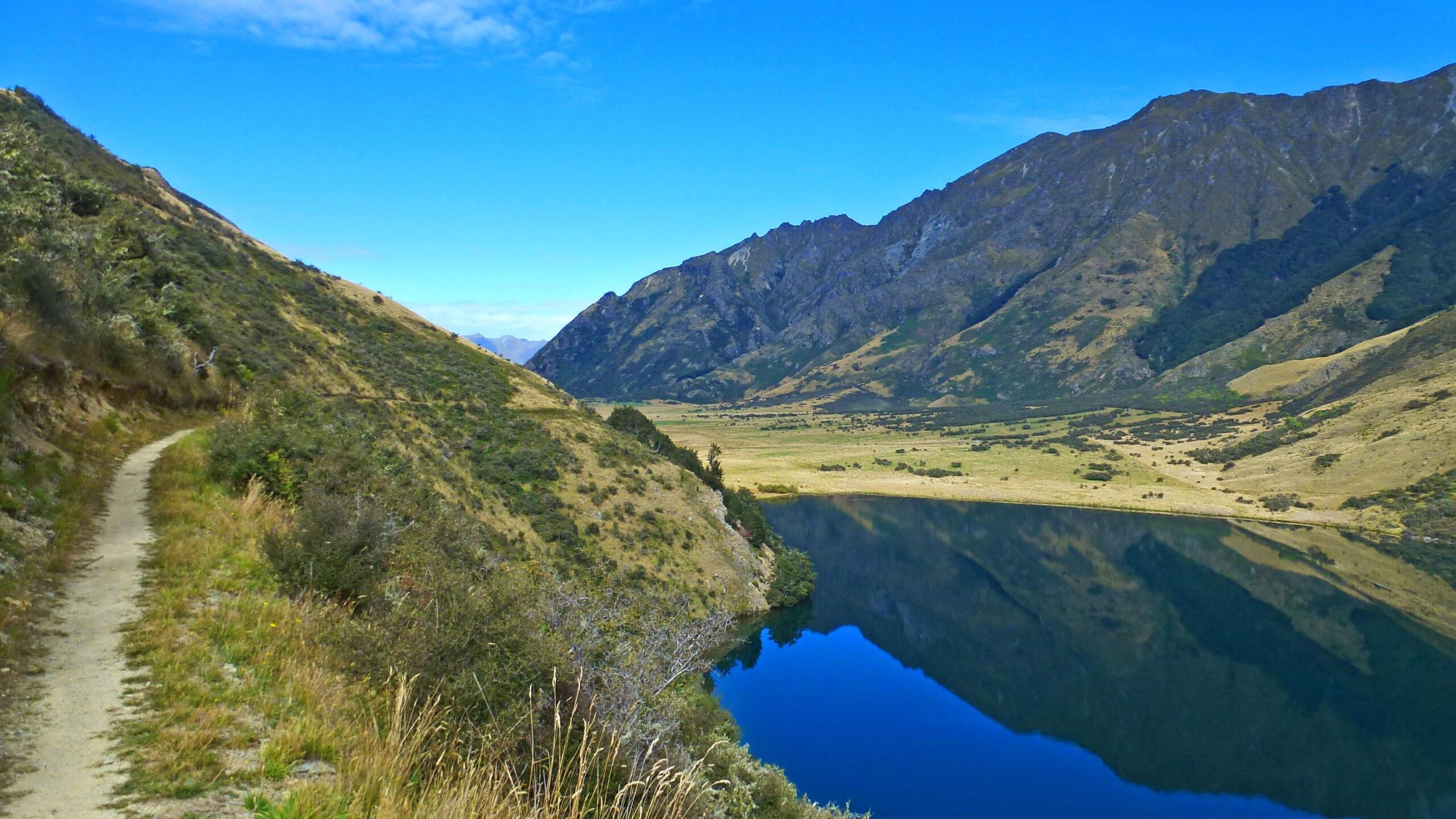 A serene landscape featuring a winding dirt path alongside a calm lake, surrounded by rolling hills and mountains. The clear blue sky reflects on the water
