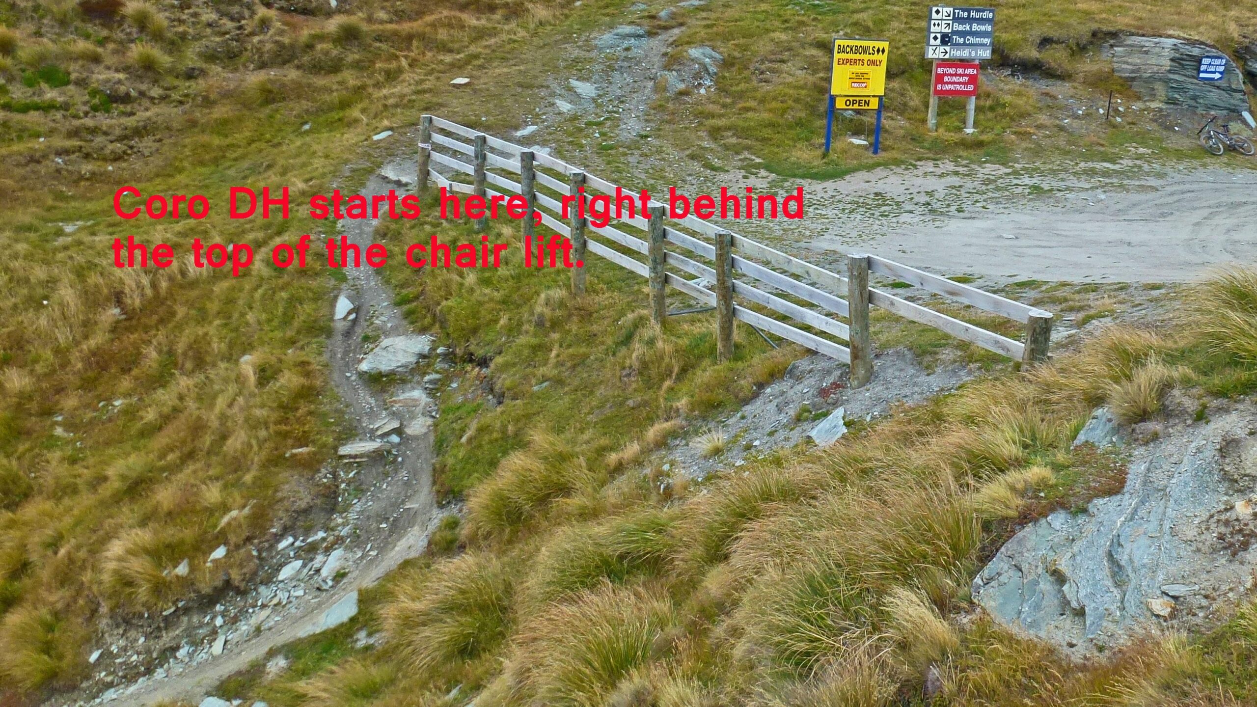 A trail entrance marked by a wooden fence, indicating the start of the Coro DH bike path, located near the top of a chair lift. Surrounding the area are grassy slopes and various signs indicating trails and expert zones. Coro DH mountain bike trail.