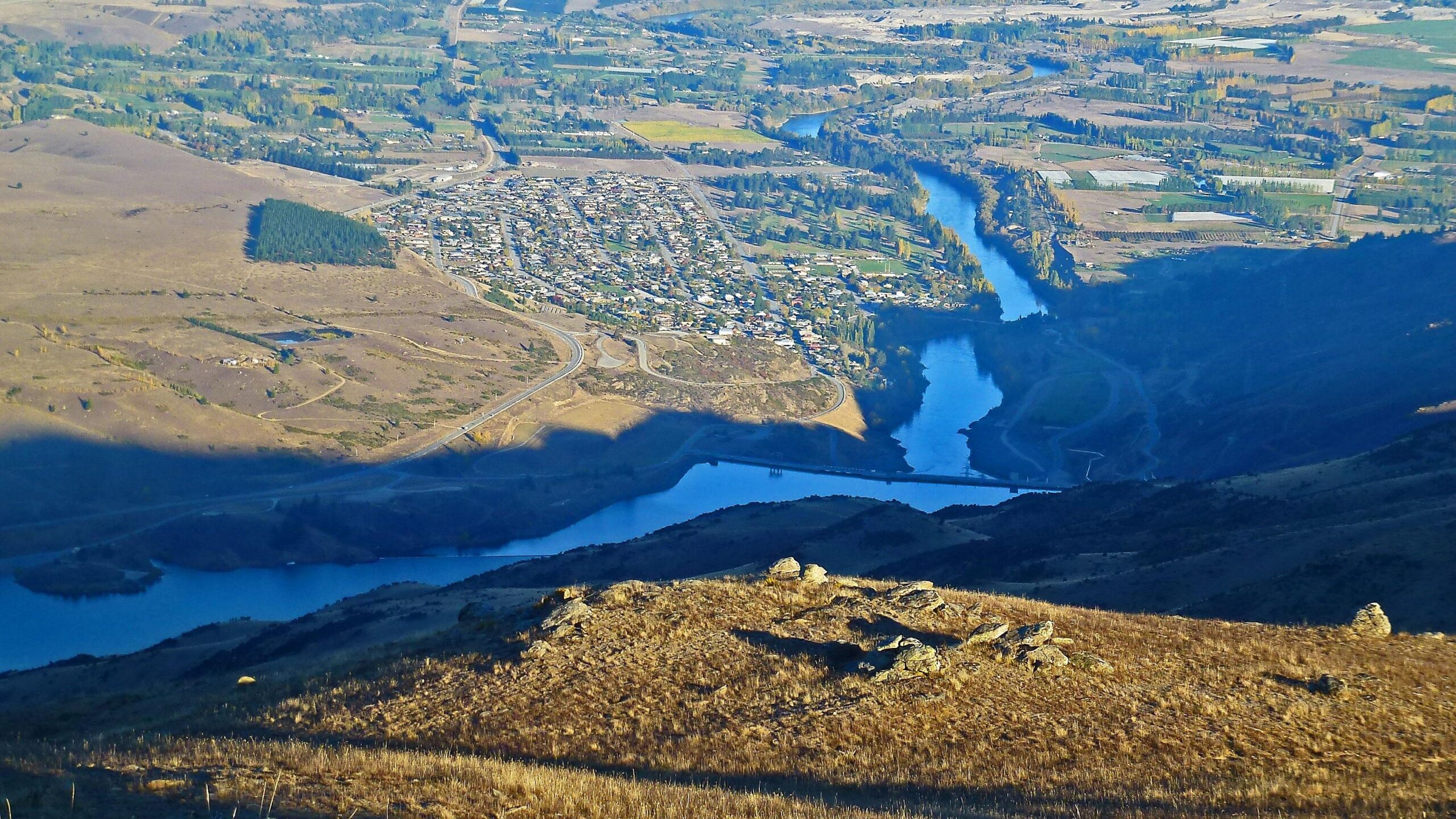 Aerial view of a river winding through a valley, with a nearby small town visible. The landscape features hills with dry grass and scattered rocks in the foreground. Lush green areas and fields can be seen in the distance, along with winding roads. The scene captures a mix of natural and developed land. Cairnmuir Hill Trail mountain bike trail.