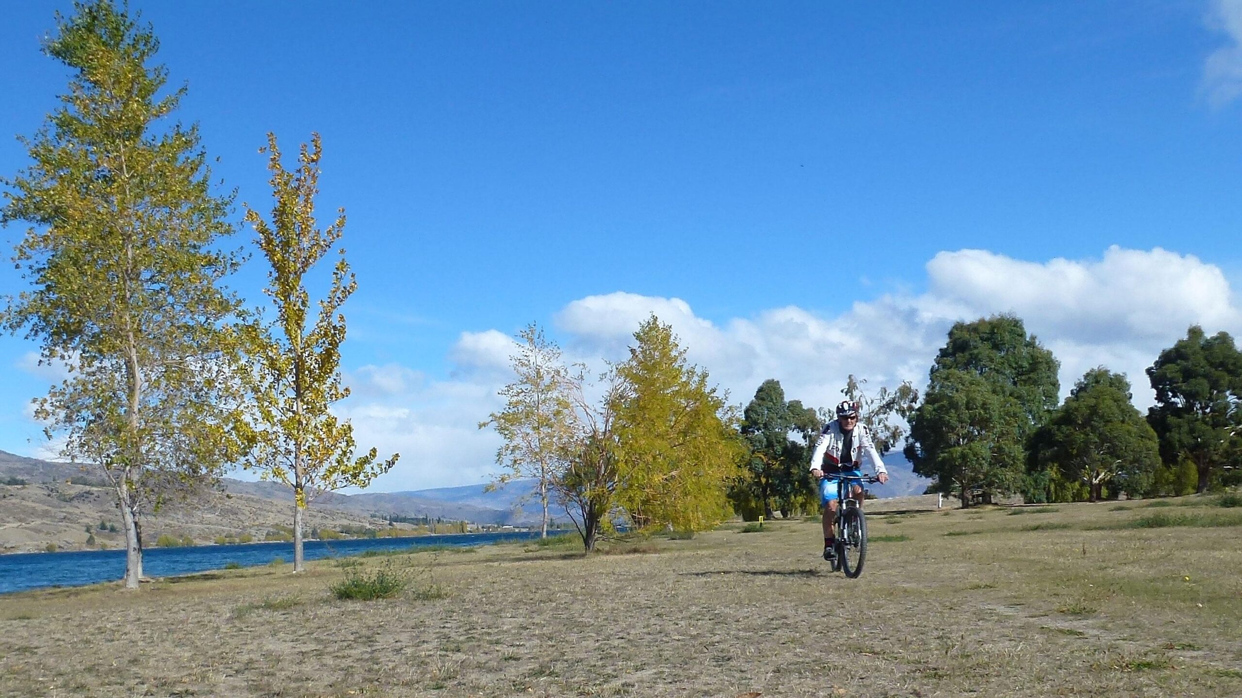 A person riding a mountain bike along a grassy pathway near a lake, surrounded by trees with autumn foliage and a clear blue sky. Dunstan Lakeside Trails mountain bike trail.