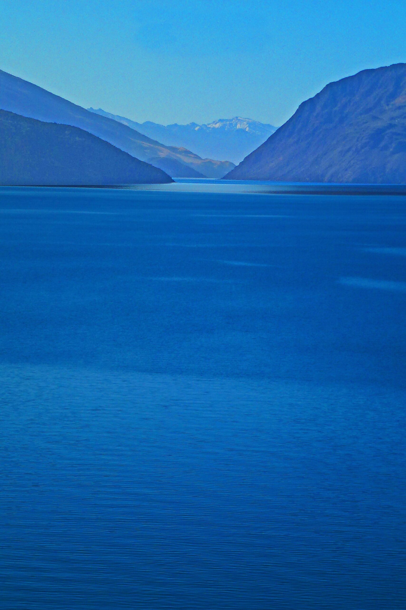 A serene landscape featuring a tranquil blue body of water surrounded by mountains. The scene showcases varying shades of blue, with distant mountains partially covered in snow, under a clear blue sky. Glendhu Bay Track mountain bike trail.