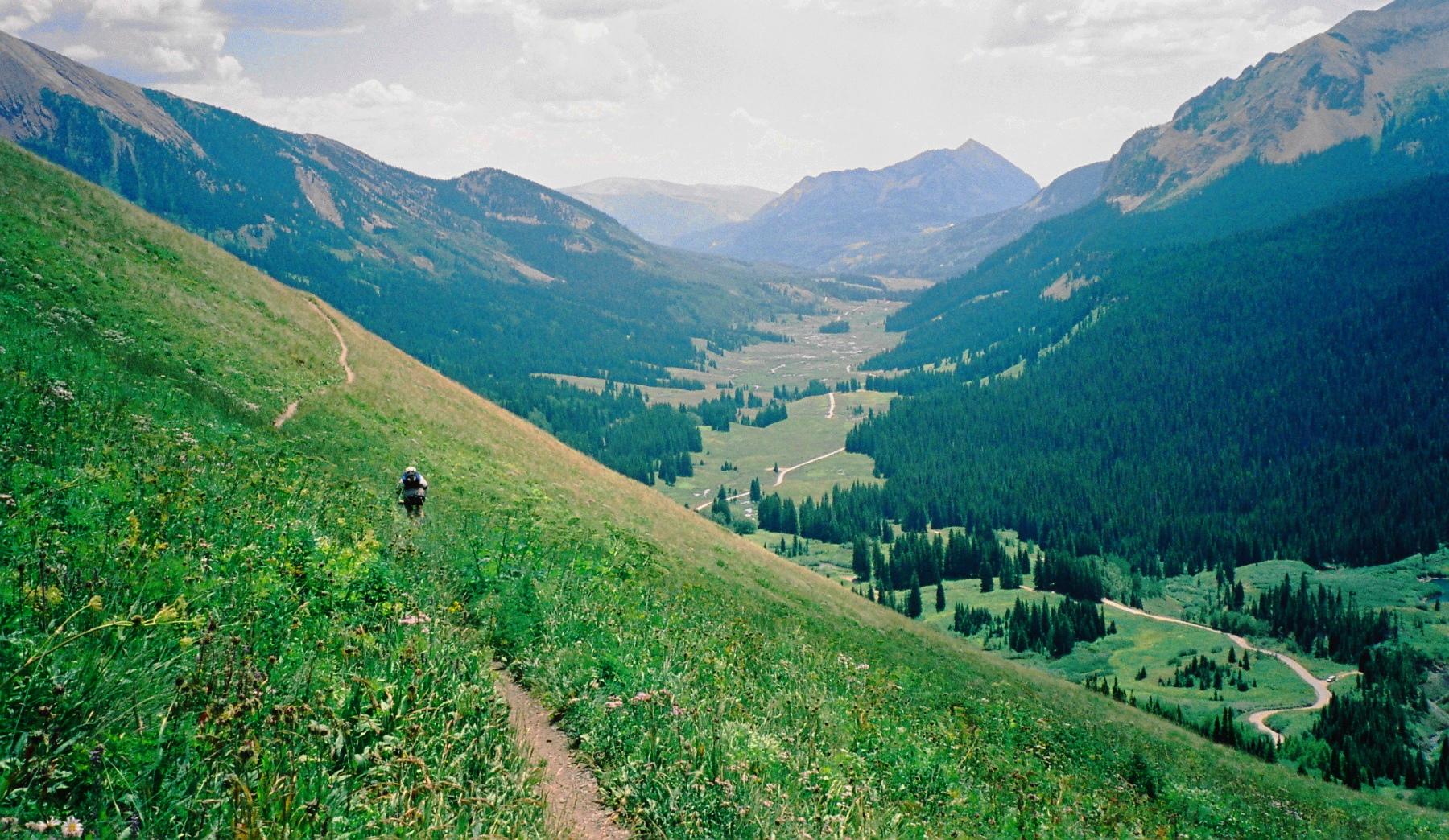 A scenic view of a mountainous landscape, featuring a hiker walking along a winding dirt trail on a grassy slope. In the background, lush green valleys and evergreen trees are visible, framed by distant mountains under a partly cloudy sky. Trail 401 mountain bike trail.