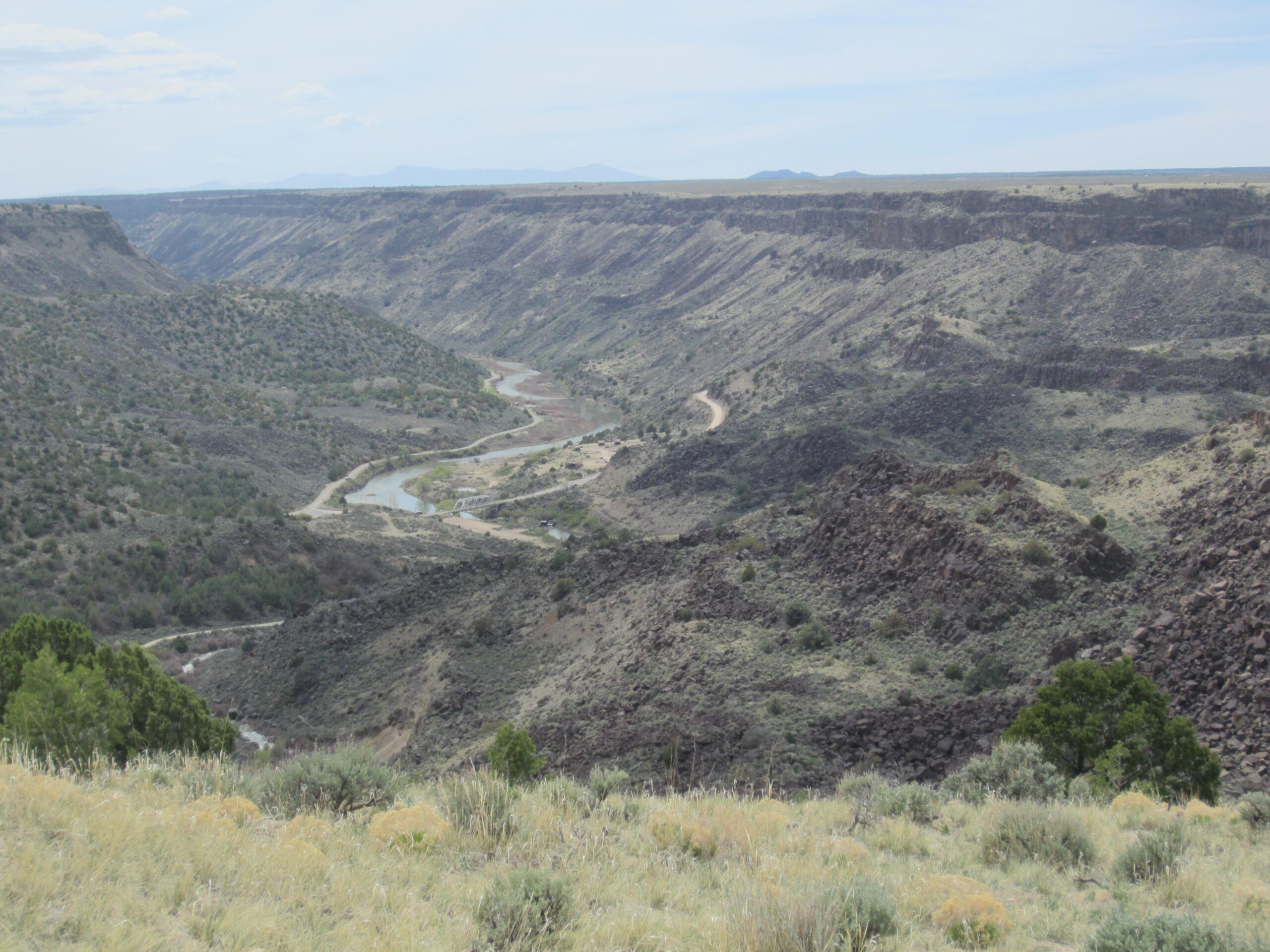 A scenic view of a deep canyon with steep, rocky cliffs and a winding river at the bottom. The landscape features sparse vegetation, with patches of green shrubs and trees, and a clear blue sky overhead. Taos Valley Overlook mountain bike trail.