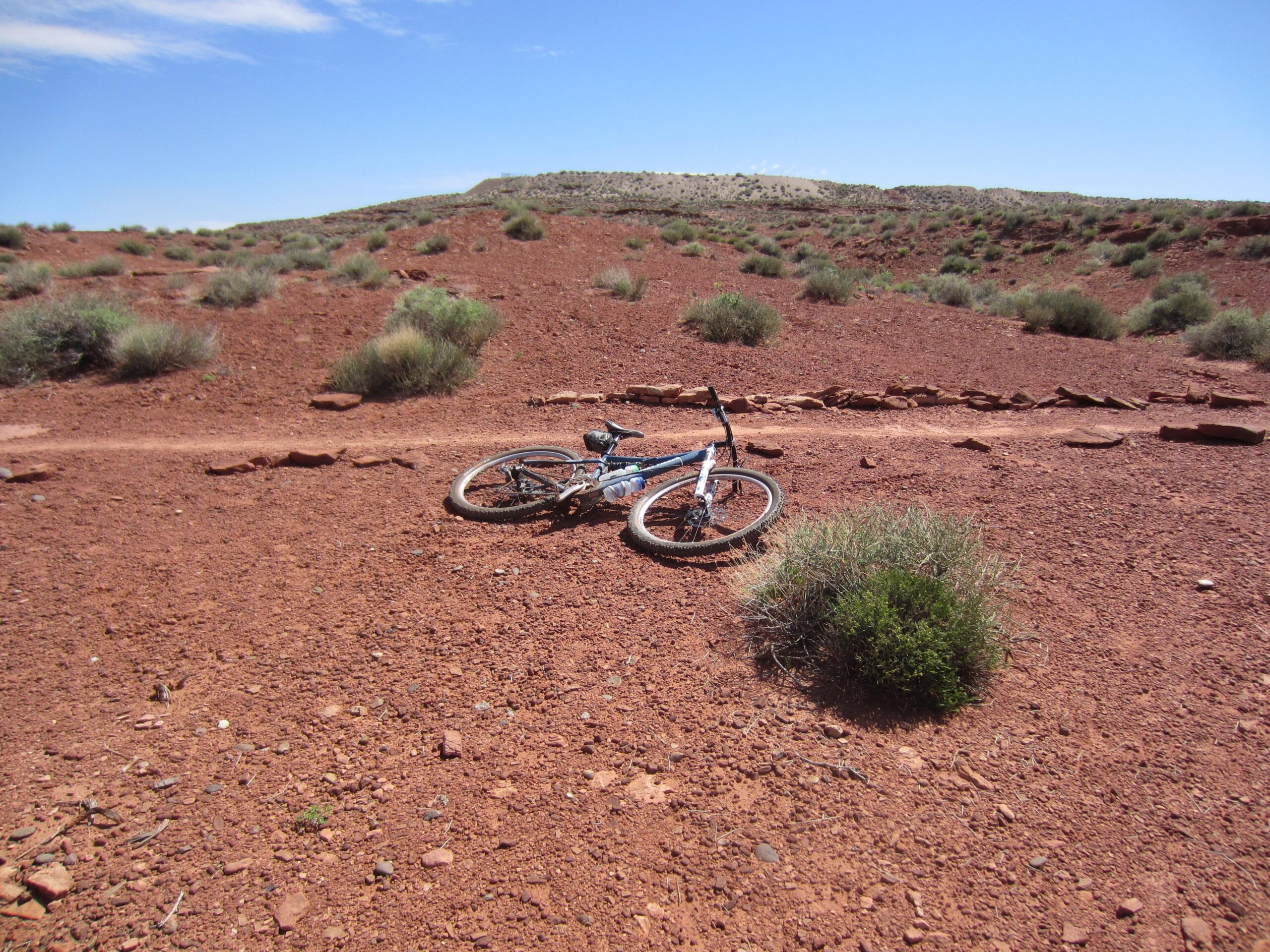 Gary Fisher HiFi Plus 29: A mountain bike lying on a reddish-brown dirt trail surrounded by sparse vegetation and rocky terrain under a clear blue sky.