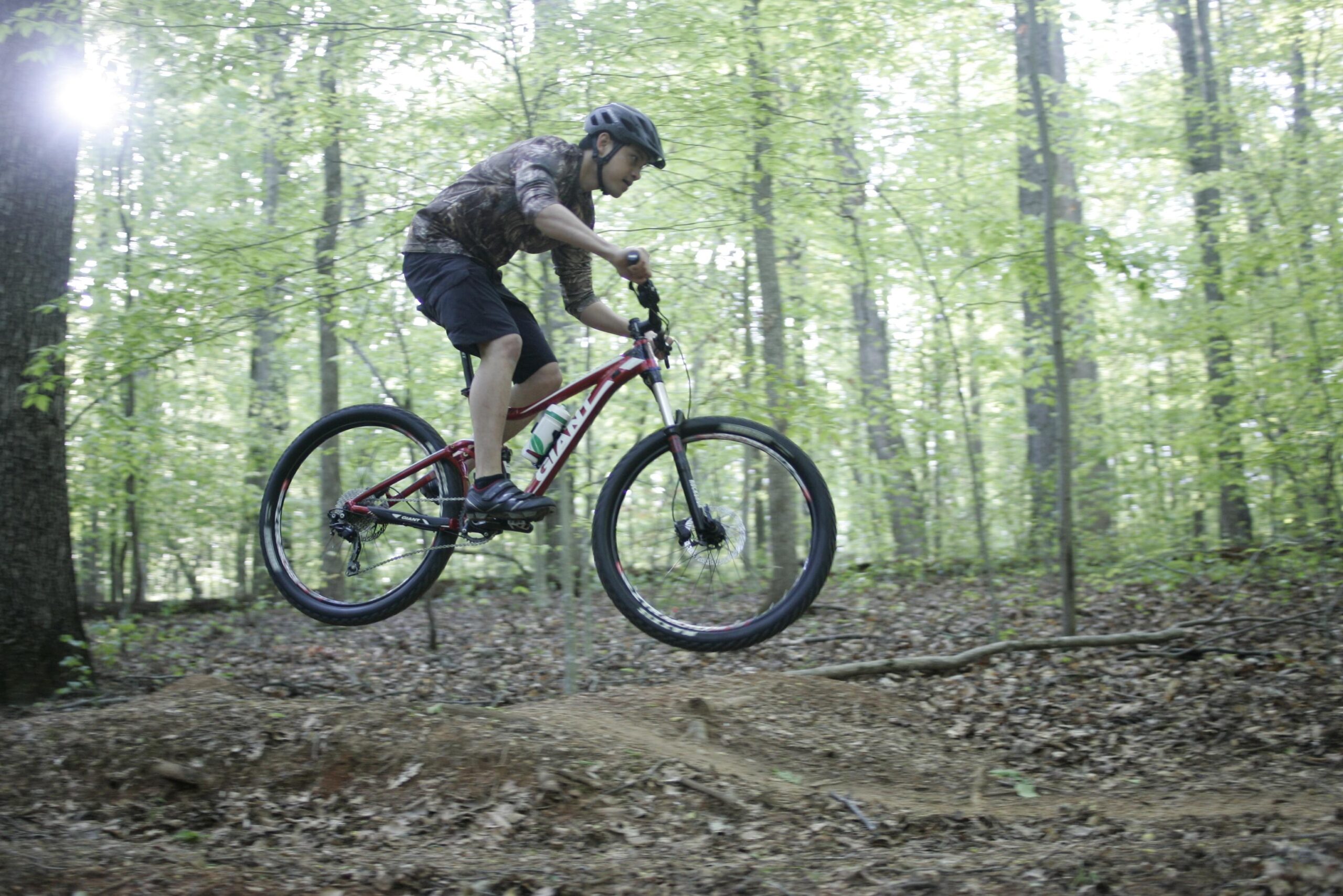 A person wearing a helmet and a camouflage shirt is mid-air while performing a jump on a mountain bike in a lush green forest. The surrounding area features trees with fresh leaves and a sunny atmosphere. Tanglewood Park mountain bike trail.