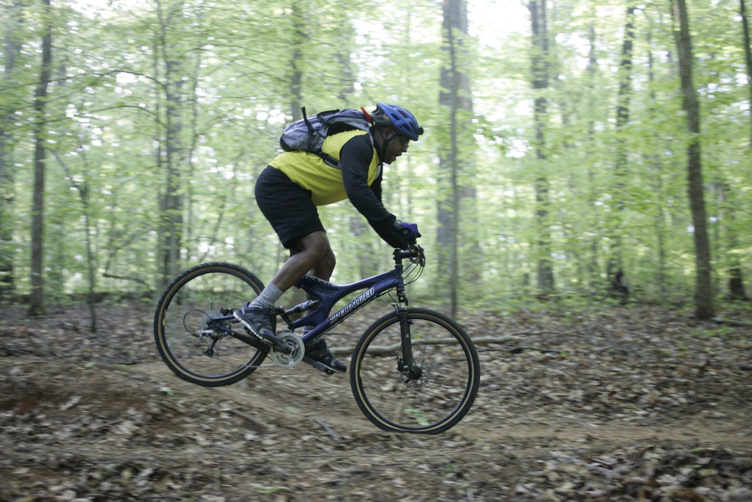 A person riding a mountain bike on a forest trail, with vibrant green trees in the background. The cyclist is wearing a blue helmet and a yellow and black shirt, positioned mid-jump on the bike. The ground is covered in leaves and dirt, indicating an outdoor adventure activity. Tanglewood Park mountain bike trail.