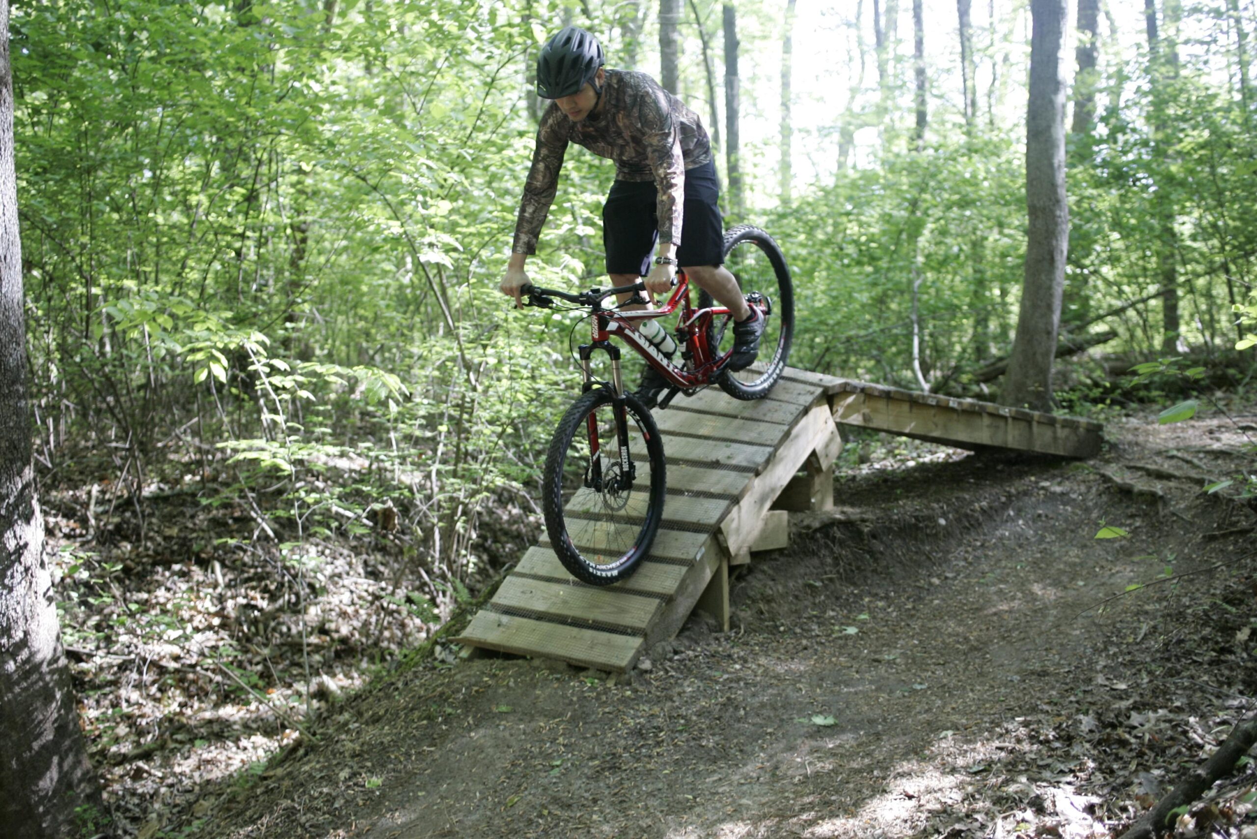 A person riding a mountain bike on a wooden ramp in a lush forest setting, with green foliage surrounding them and a dirt trail nearby. The cyclist is leaning forward, navigating the incline of the ramp. Tanglewood Park mountain bike trail.