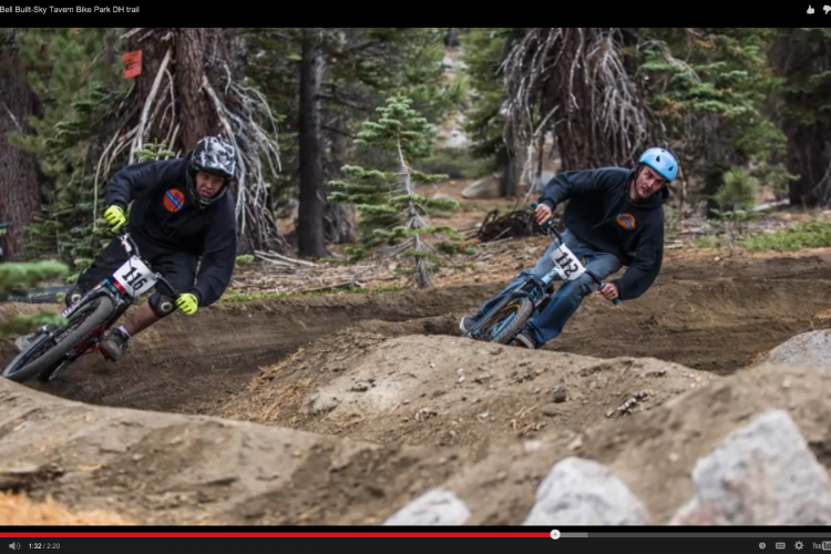 Two mountain bikers race along a dirt trail in a bike park, maneuvering through a banked turn. One rider, wearing a black hoodie and helmet, is closer to the camera, while the other, in a blue helmet and similar attire, rides slightly behind. The background features tall trees and rocky terrain, emphasizing the outdoor setting of the bike park.
