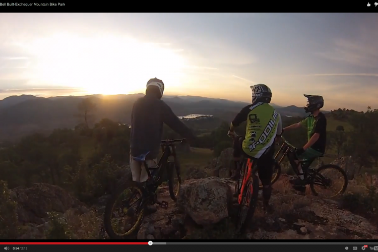 Three mountain bikers are standing on a rocky outcrop, overlooking a scenic landscape during sunset. The sun casts a warm glow over the mountains in the background, with a lake visible in the distance. Two riders are wearing helmets and jerseys, one in green with black and white accents, and the other in dark clothing. The third rider, wearing a black and white helmet, is partially turned away, looking out at the view. The scene captures a moment of camaraderie and adventure in nature.