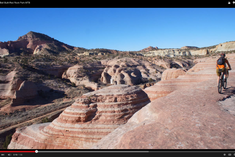 A mountain biker rides along a rocky trail in Bell Built-Red Rock Park, featuring striking layered rock formations and a clear blue sky in the background. The landscape includes various shades of red and tan from the terrain and unique geological structures.