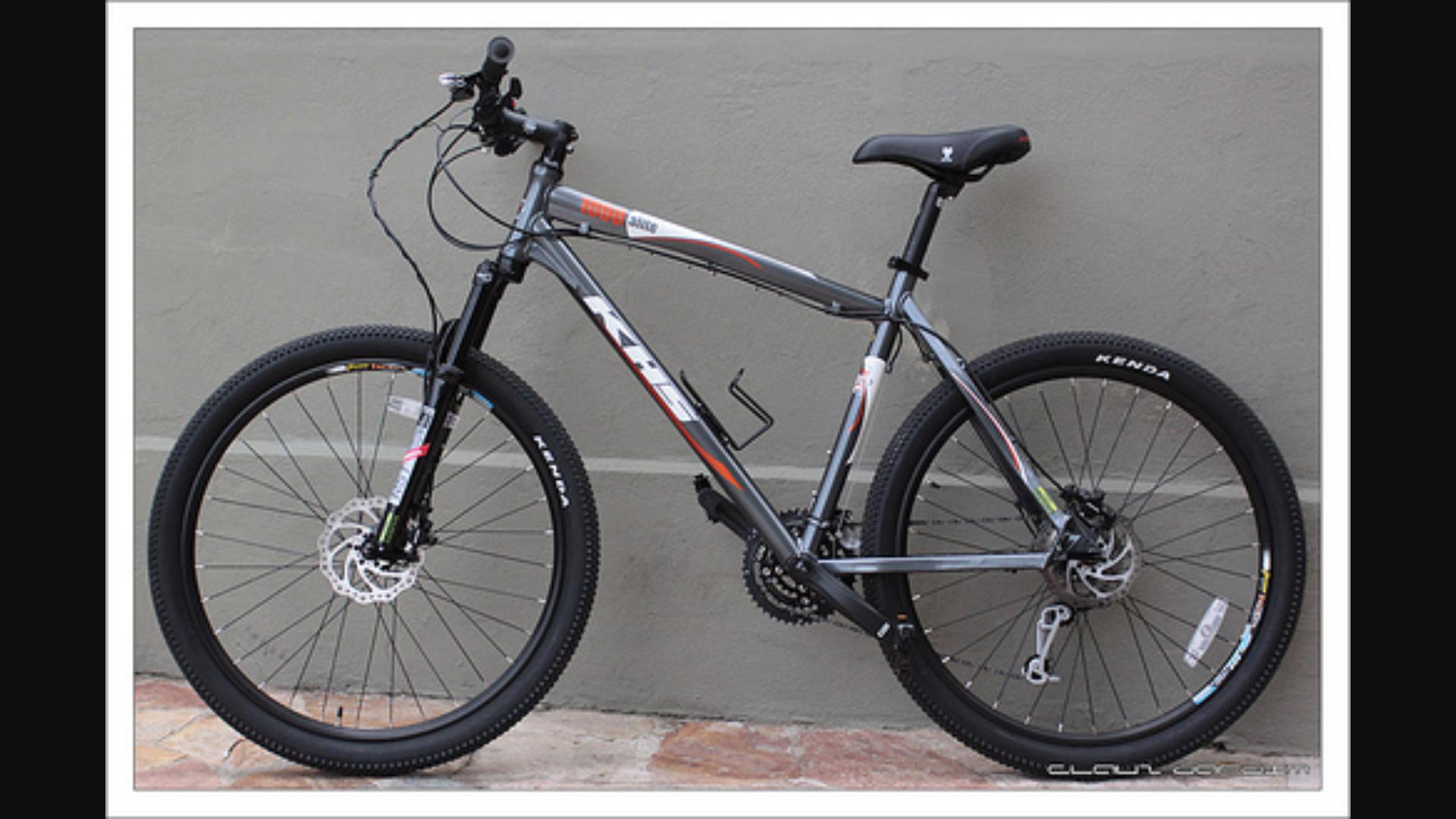 KHS Alite 1000: A gray mountain bike with black tires and disc brakes, featuring a sleek frame design, adjustable seat, and textured handle grips, parked against a plain wall.