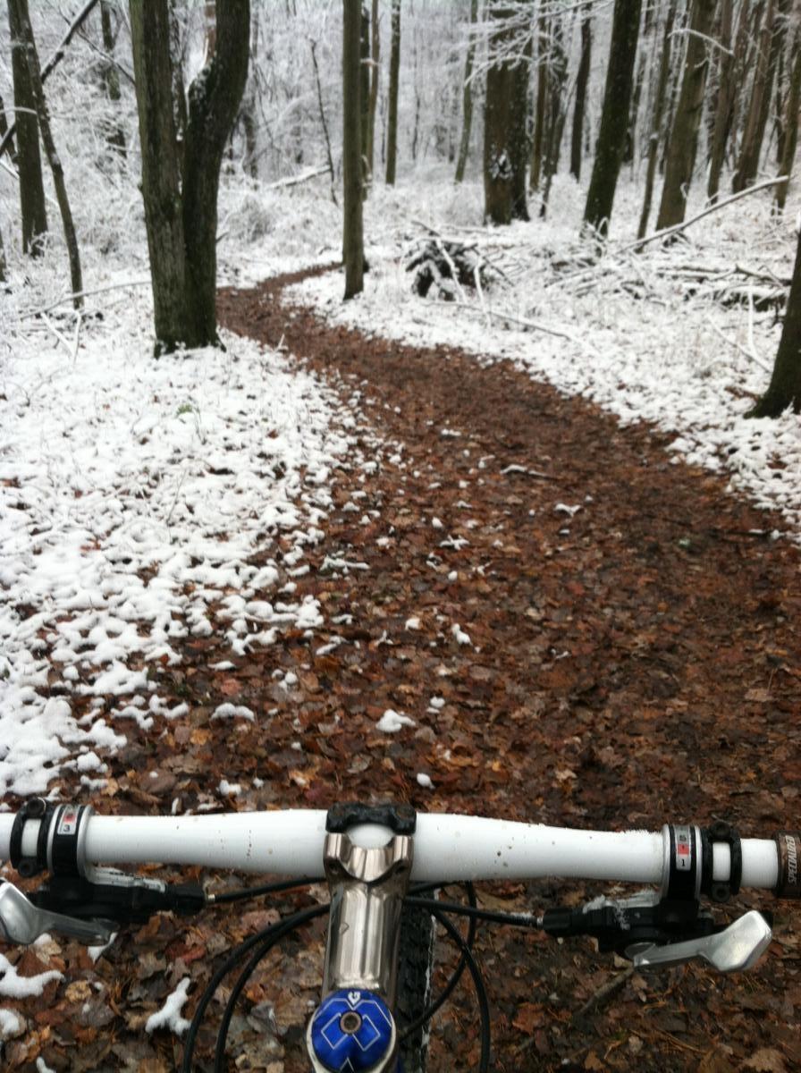 A snowy forest trail seen from the perspective of a mountain bike handlebars, with a winding path covered in fallen leaves and patches of snow on the ground. Trees line the sides of the trail, creating a serene winter atmosphere. Mountwood mountain bike trail.