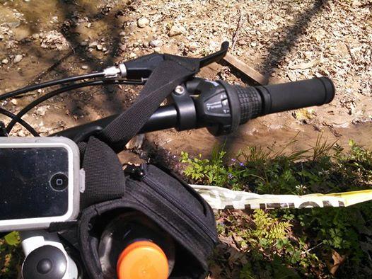 Alt text: Close-up of a bicycle handlebar featuring a smartphone mounted in a holder, a black grip shifter, and a water bottle in a pouch, with a rocky creek and vegetation in the background. Sockum Ridge mountain bike trail.