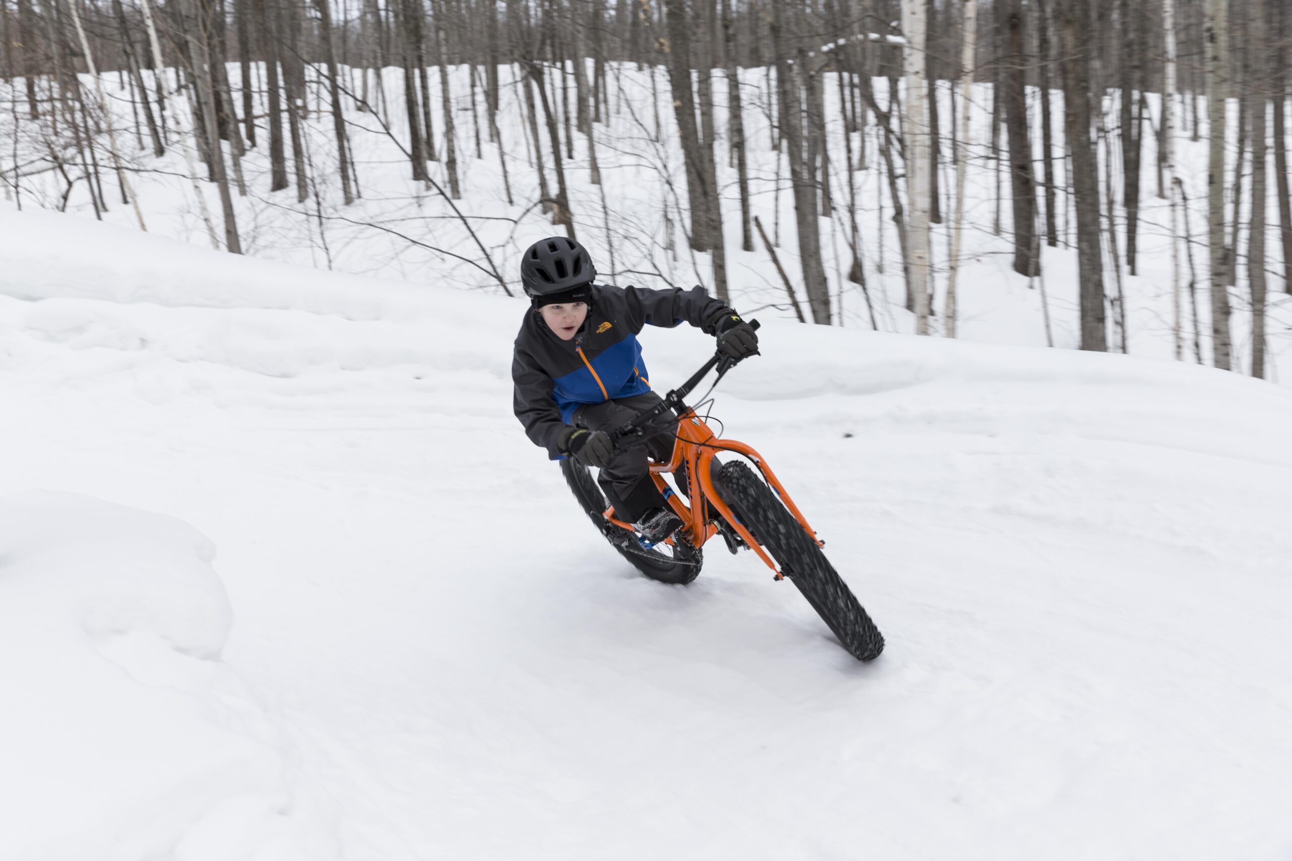 Trek Farley: A child riding a fat tire bike on a snow-covered path, leaning into a turn. The background features a forest of bare trees, suggesting a winter setting. The child is wearing a helmet and winter clothing with a blue and black jacket.