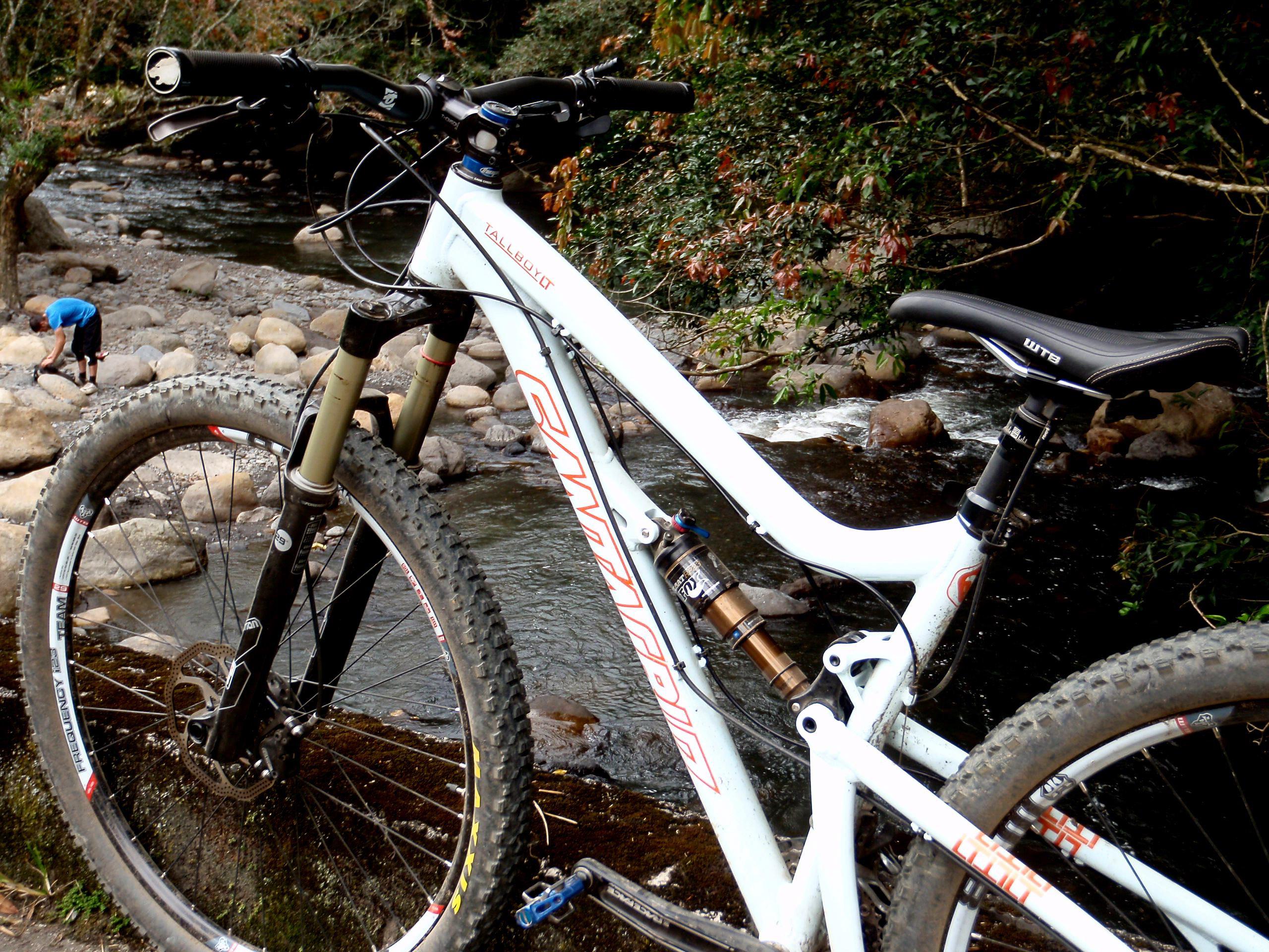 Santa Cruz Tallboy LT: A close-up view of a mountain bike parked on the edge of a rocky riverbank, with a flowing stream and lush greenery in the background. A person in the distance is crouched down near the water, surrounded by smooth stones. The bike features thick tires and a prominent suspension system.