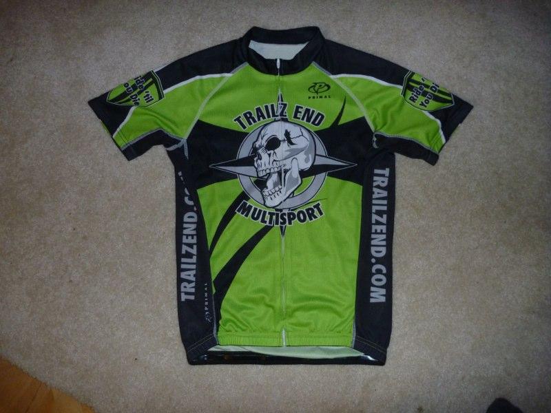 A close-up view of a short-sleeved cycling jersey featuring a bright green and black design. The jersey prominently displays the logo of "Trailz End Multisport," which includes a graphic of a skull with crossed arrows. Additional branding is visible on the sleeves and sides, emphasizing a sporty and adventurous aesthetic. The jersey is laid flat on a light-colored carpet background.