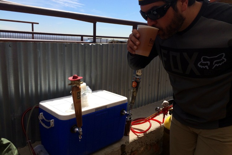 A man in sunglasses and a black long-sleeve shirt stands in front of a cooler with a beer tap, holding a cup and taking a sip. The backdrop features a clear blue sky and a railing, suggesting an outdoor setting. The cooler is connected to hoses, and a small decorative figure with a sombrero is perched on the beer tap.