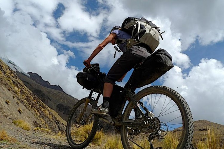 A person riding a mountain bike uphill on a rocky terrain, dressed in outdoor gear and carrying a large backpack. The scene features a dramatic sky with clouds and distant mountain peaks in the background.