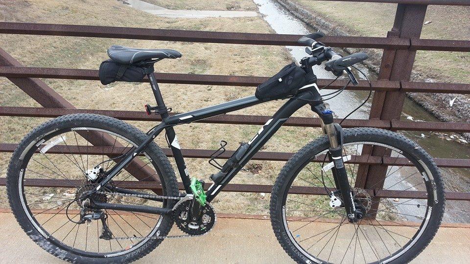 Trek Mamba Gary Fisher: A black mountain bike resting on a bridge with a grassy area and a small stream in the background. The bike features thick tires, a small saddlebag on the seat, and a sturdy frame. The setting is outdoors, suggesting a scenic location for cycling.