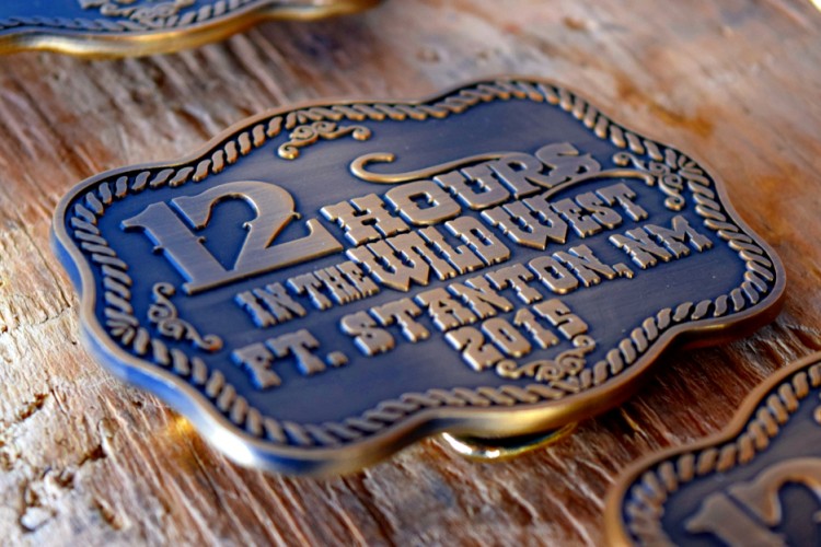 A bronze medal featuring decorative text that reads "12 HOURS IN THE WILD WEST, FT. STANTON, NM, 2015." The medal is artistically designed with rope-like borders and is resting on a rustic wooden surface.