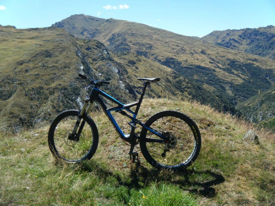 Specialized Enduro Expert Carbon: A mountain bike resting on a grassy hilltop, with a panoramic view of rolling hills and mountains in the background under a clear blue sky.
