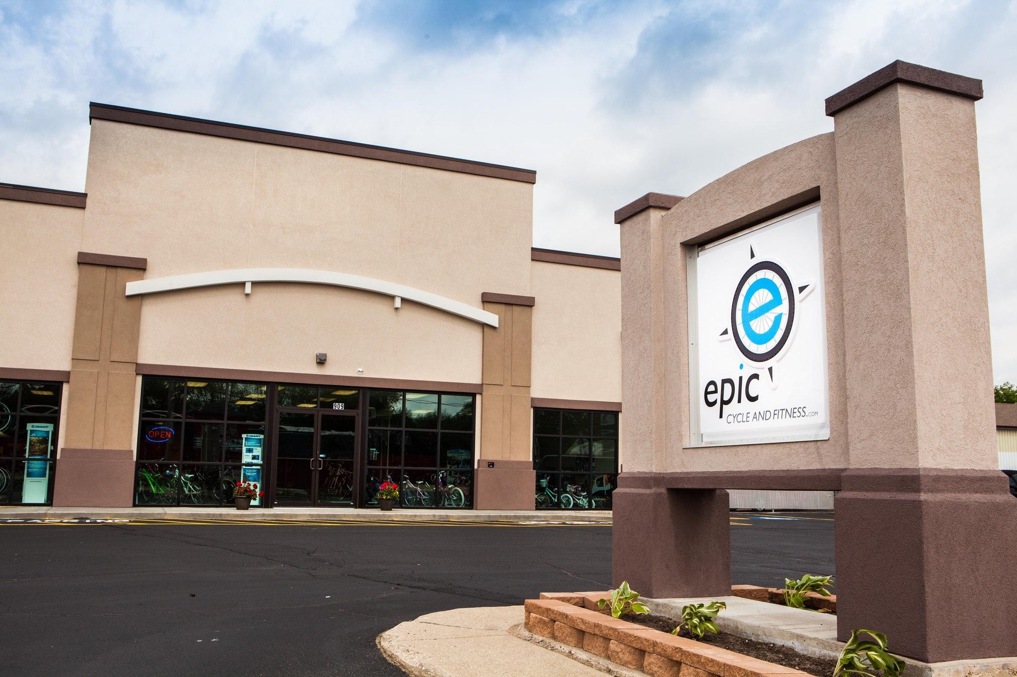 Image of the exterior of a business named "Epic Cycle and Fitness." The building features large windows displaying bicycles inside, with a sign out front prominently displaying the business name. The area is paved, with planters at the base of the sign and a cloudy sky above.