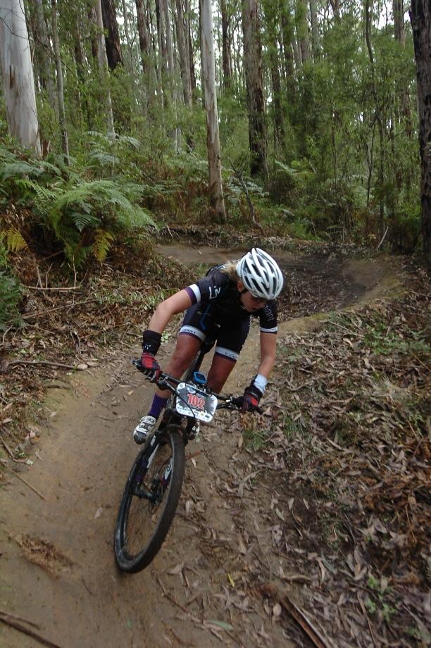 A mountain biker navigating a narrow dirt trail in a forested area, wearing a helmet and cycling gear. The cyclist leans forward on the bike, concentrating on the path ahead, surrounded by tall trees and ferns. Forrest Mtb Trails mountain bike trail.