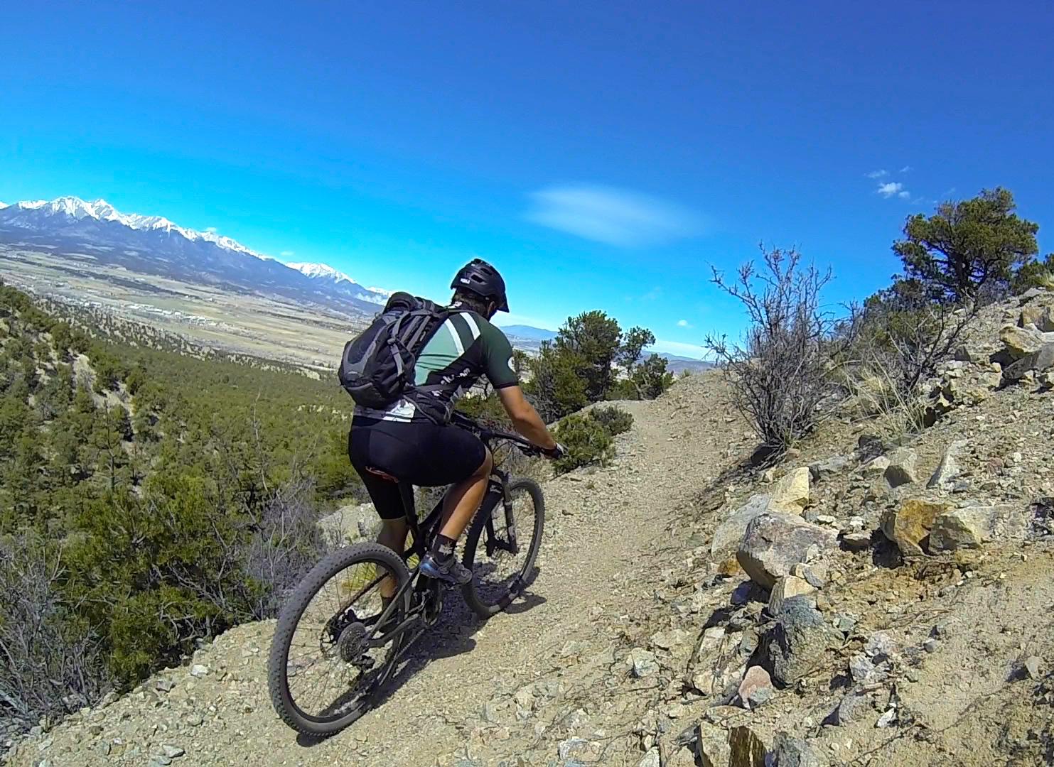 A person riding a mountain bike along a narrow dirt trail, surrounded by rocky terrain and sparse vegetation. In the background, a panoramic view of snow-capped mountains under a clear blue sky can be seen. Double Rainbow mountain bike trail.