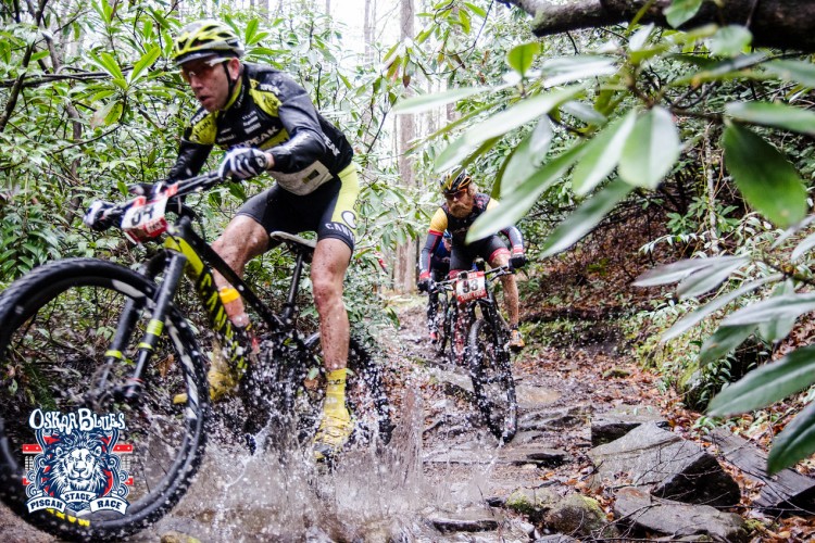 Two mountain bikers navigate a rocky, muddy trail surrounded by lush green foliage. The rider in the foreground splashes through a puddle, demonstrating speed and skill. Both cyclists wear competitive gear with identification numbers and logos, indicating participation in a race. The image captures the intensity and excitement of outdoor racing in a natural setting.