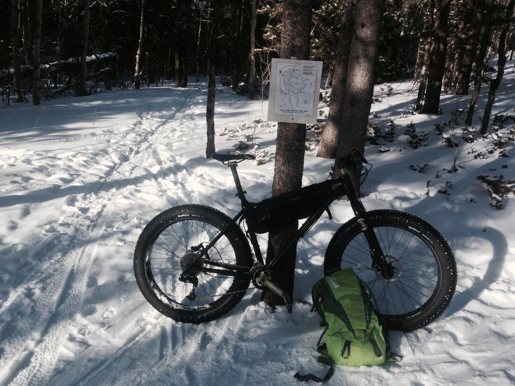 A fat bike resting on a snowy trail, next to a tree with a trail map attached. A green backpack is placed on the snow near the bike, surrounded by a winter landscape of snow-covered ground and trees. Happy Jack Recreation Area mountain bike trail.