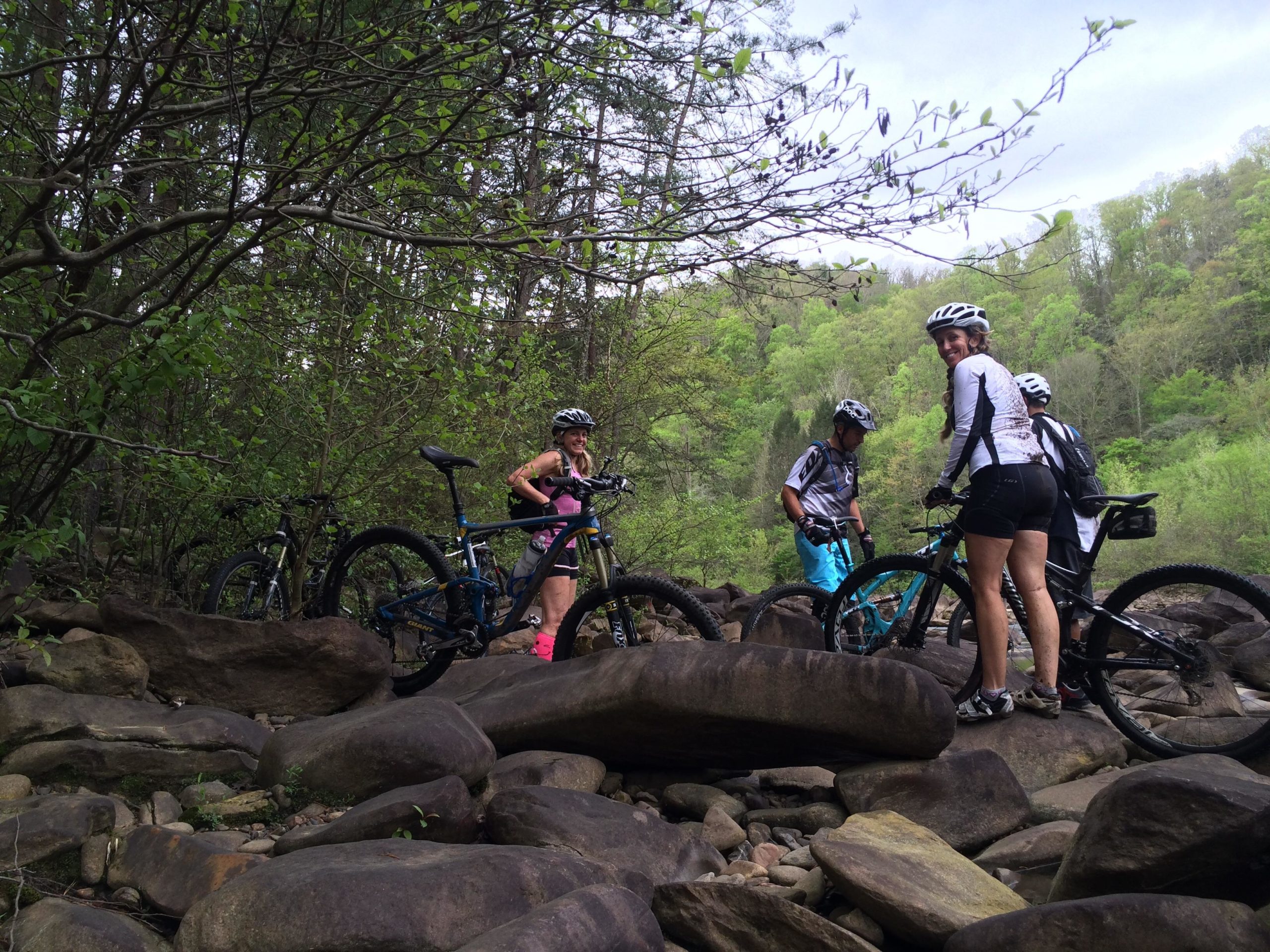 A group of four mountain bikers exploring a rocky trail surrounded by lush greenery. Two bikes are parked on the rocks, and the riders are wearing helmets and cycling gear. The background features trees and hills, capturing the serene outdoor environment. Brush Creek mountain bike trail.