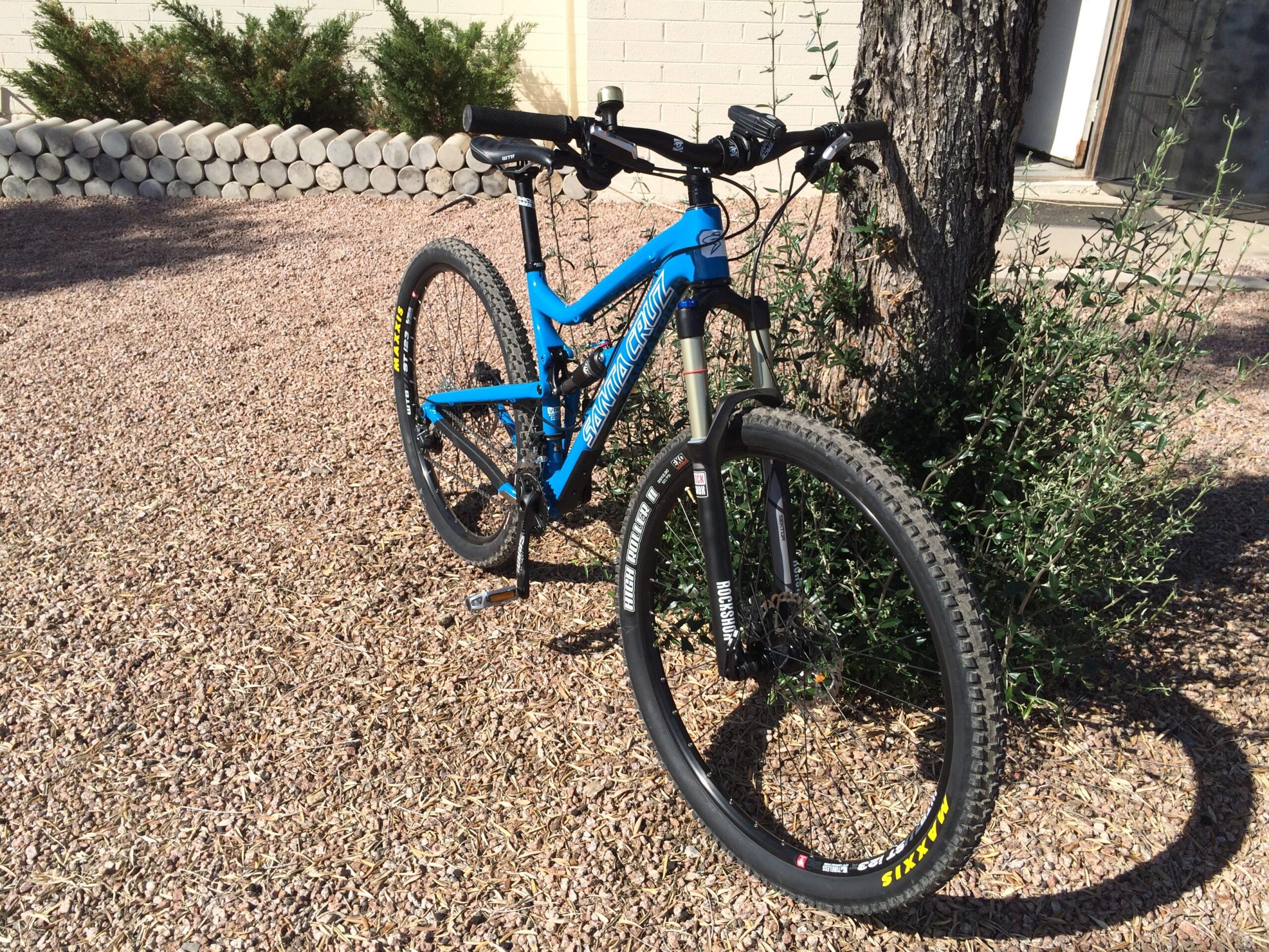 Santa Cruz Tallboy Carbon: A blue mountain bike is parked on a gravel surface near a tree and some shrubs. The bike features thick tires designed for off-road terrain and a sturdy frame, highlighting its suitability for outdoor cycling adventures.