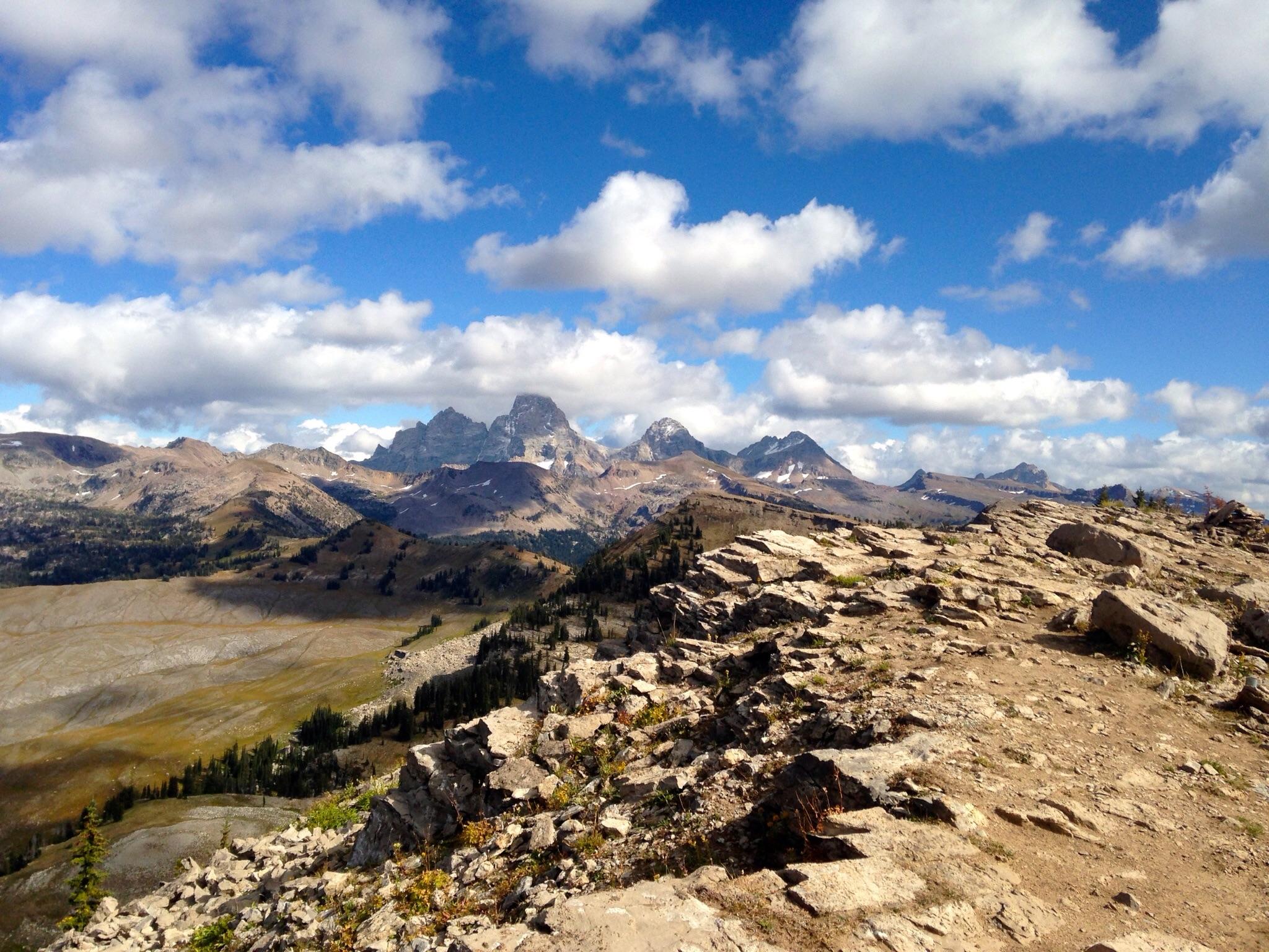 A panoramic view of rugged mountains under a partly cloudy sky. The foreground features rocky terrain and patches of grass, while the background showcases a range of peaks, some with snowcaps, surrounded by rolling hills and forests. The scene captures the beauty of a natural landscape, highlighting the interplay of light and shadow across the terrain. Grand Targhee Bike Park mountain bike trail.