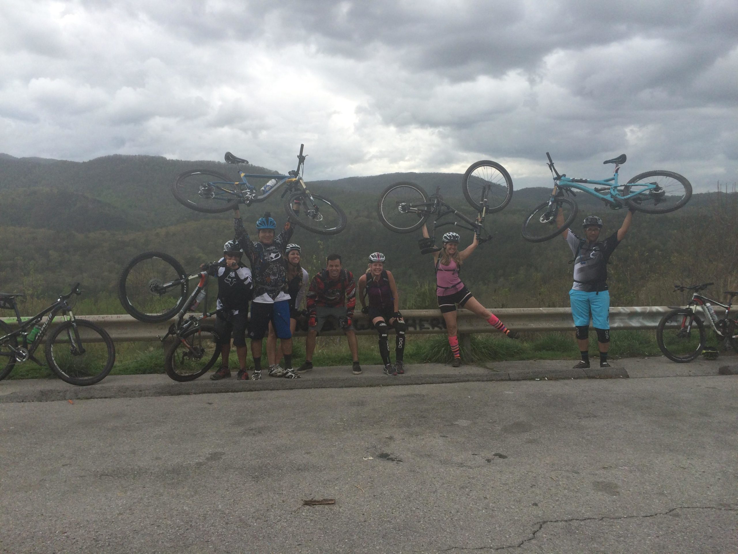 A group of nine mountain bikers posing outdoors, holding their bikes above their heads in celebration. They are standing near a roadside, with a backdrop of lush green hills and a cloudy sky. Some bikers are dressed in cycling gear and helmets, while others are wearing casual attire. Several bicycles are parked beside them. Brush Creek mountain bike trail.