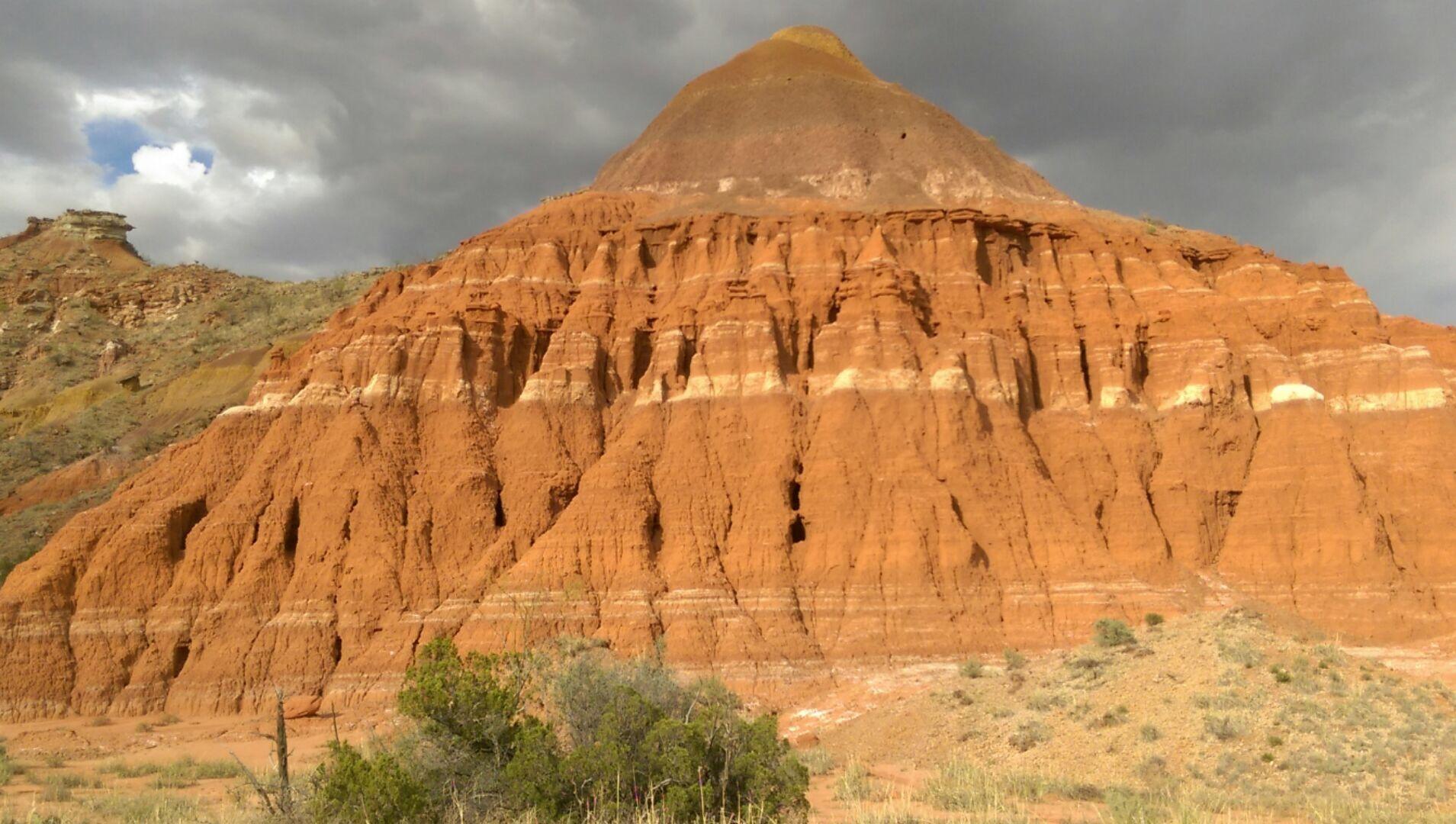 A dramatic landscape featuring a prominent reddish-brown hill with layered textures, under a cloudy sky. The hill has a rounded peak and is surrounded by arid terrain with sparse vegetation. Palo Duro Canyon mountain bike trail.
