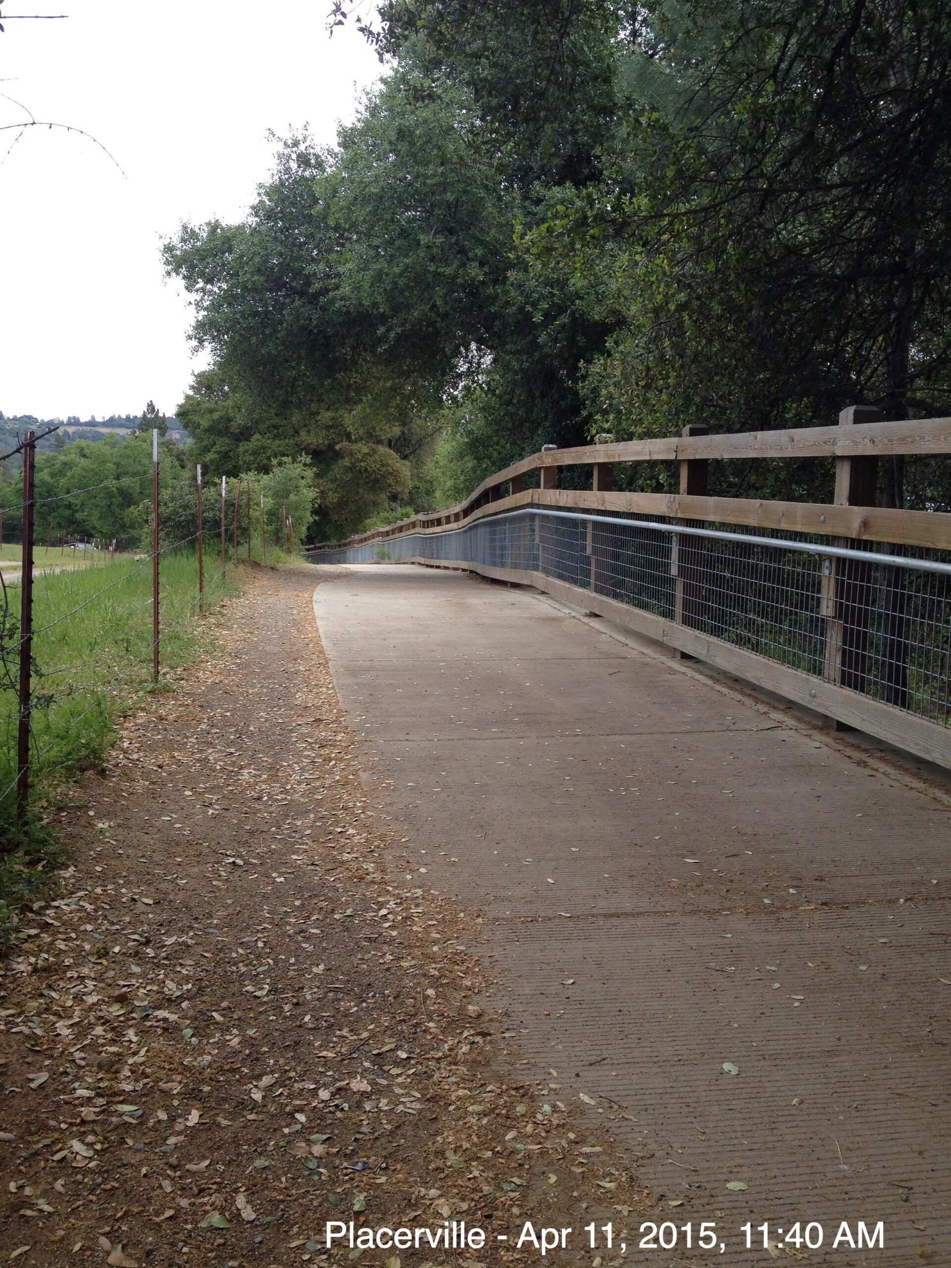 A winding paved path bordered by grass and trees, with a wooden railing on one side. The scene is serene, capturing a tranquil outdoor area, possibly in a park or nature reserve. Foliage covers the ground, hinting at a peaceful atmosphere. The image is timestamped "Placerville - Apr 11, 2015, 11:40 AM." El Dorado Trail mountain bike trail.