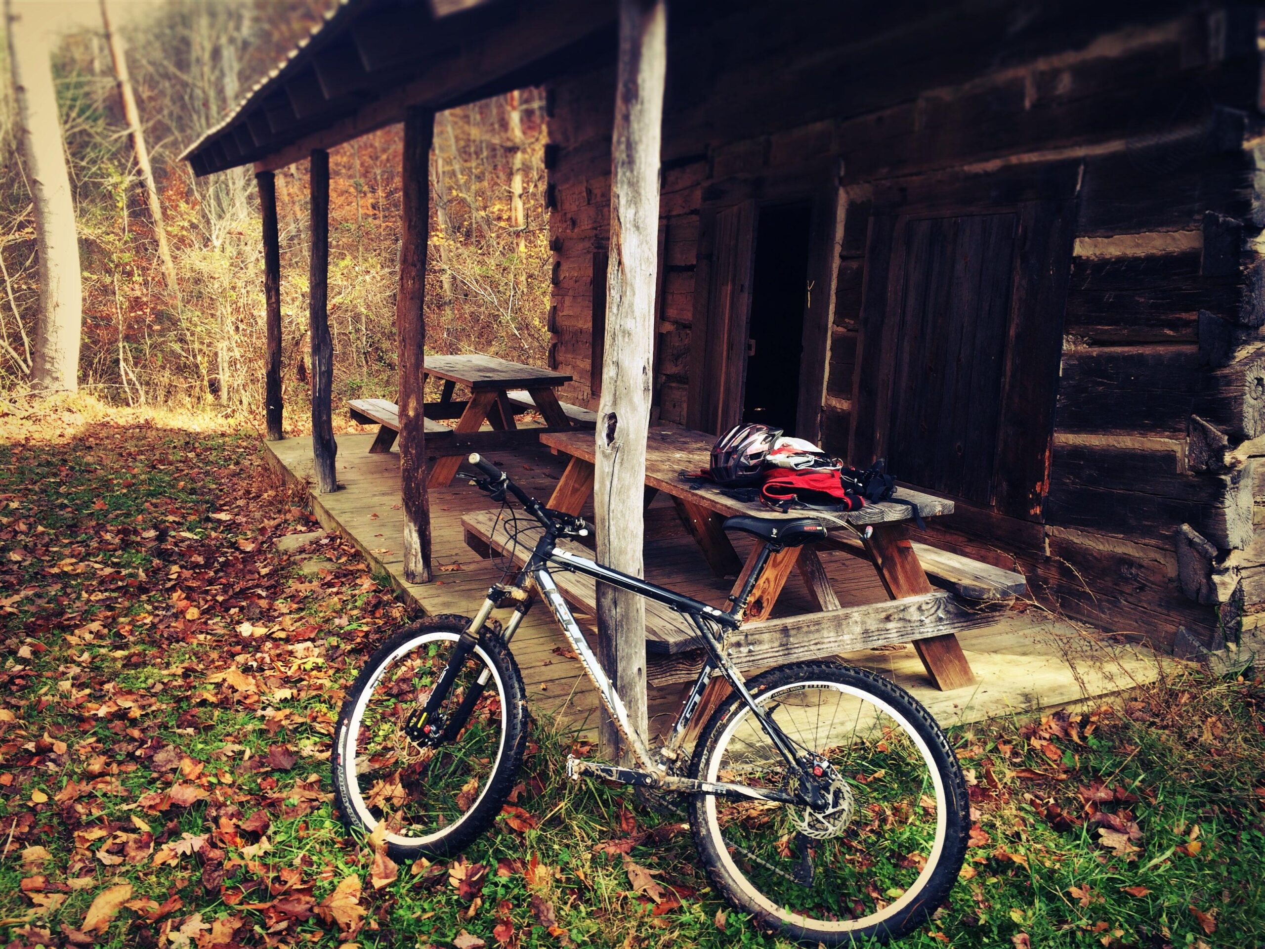 GT Avalanche: A mountain bike leaning against a wooden pillar, with a rustic log cabin in the background. A picnic table is situated on the porch, surrounded by fallen leaves on the ground and trees in autumn colors. A helmet and backpack are placed on the table.