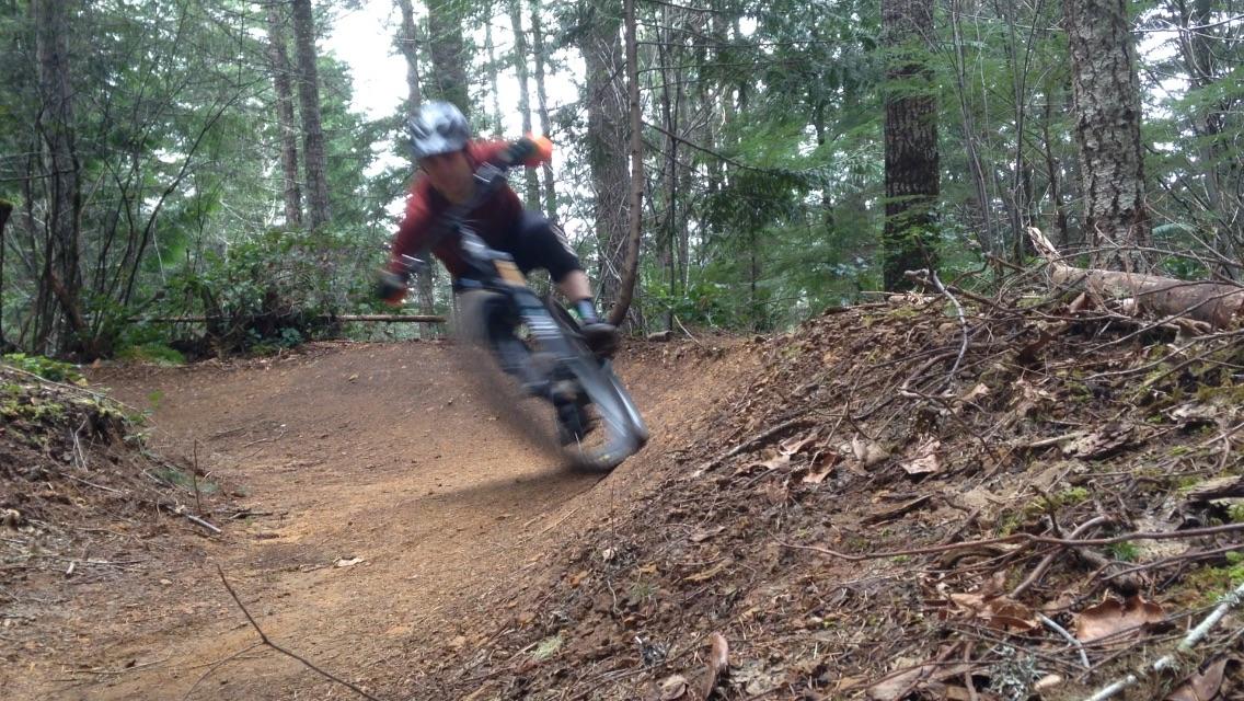 A mountain biker quickly navigating a dirt trail in a forested area, leaning into a turn, with trees and foliage visible in the background. The scene conveys the speed and excitement of off-road cycling. Alsea Falls Recreation Site mountain bike trail.