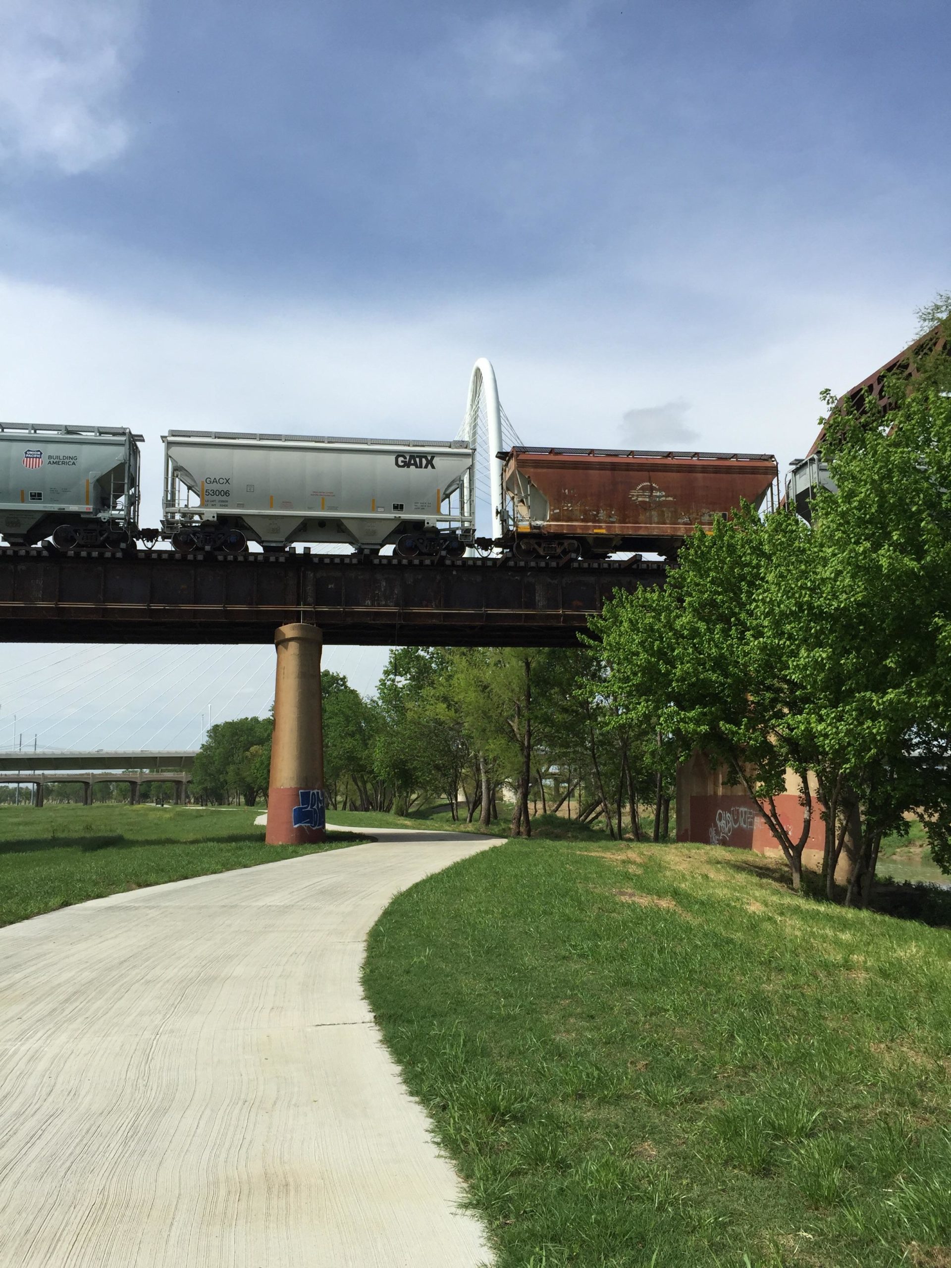 A view of a train bridge with several freight train cars passing overhead. Below, a paved path winds through a green park area with grass and trees. In the background, a modern bridge structure is visible against a partly cloudy sky. Trinity Levee mountain bike trail.