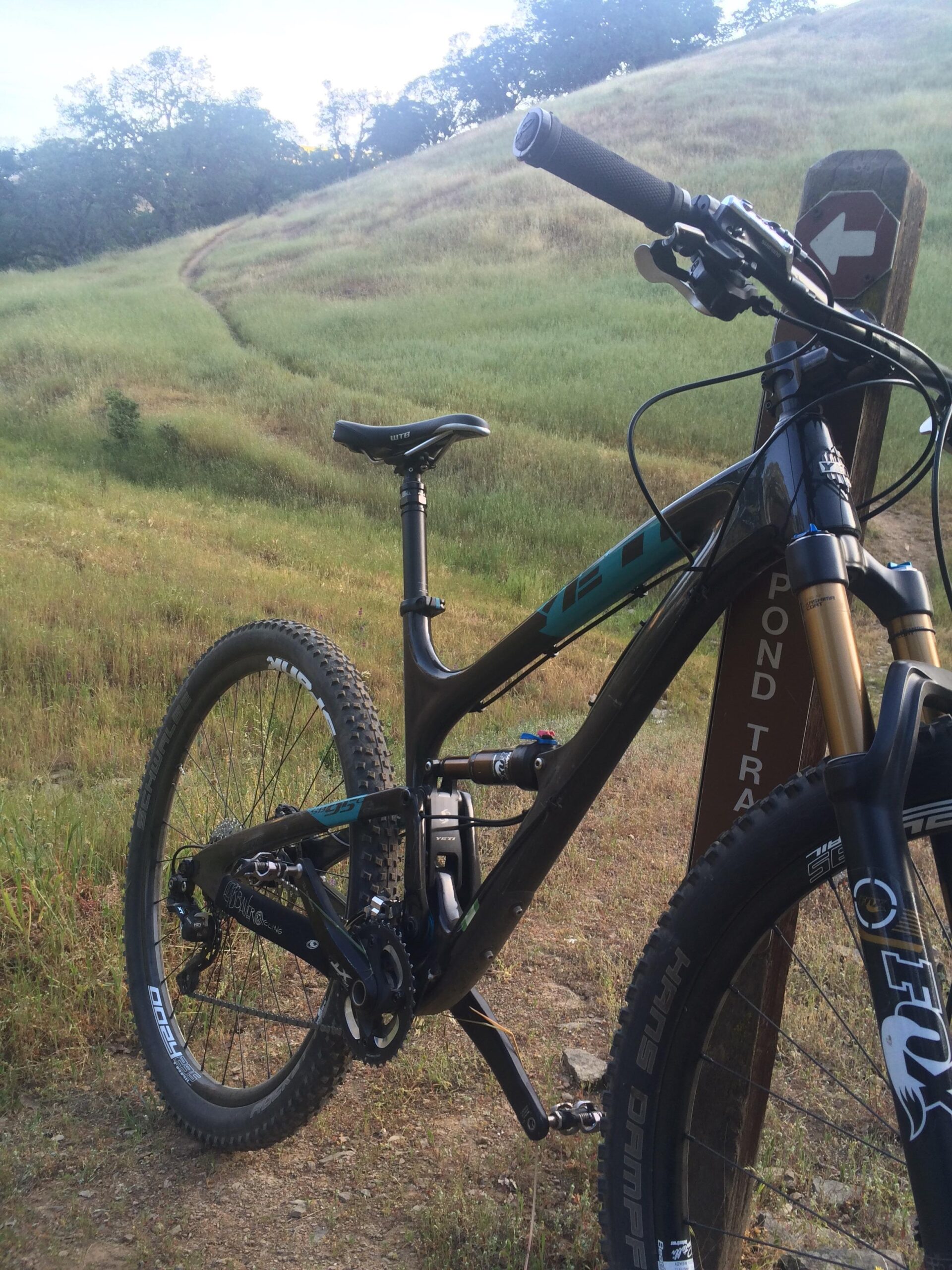 Yeti SB95: A mountain bike resting on a gravel path, next to a wooden trail sign indicating the direction to the pond trail. The background features a lush, grassy hillside under a clear sky.