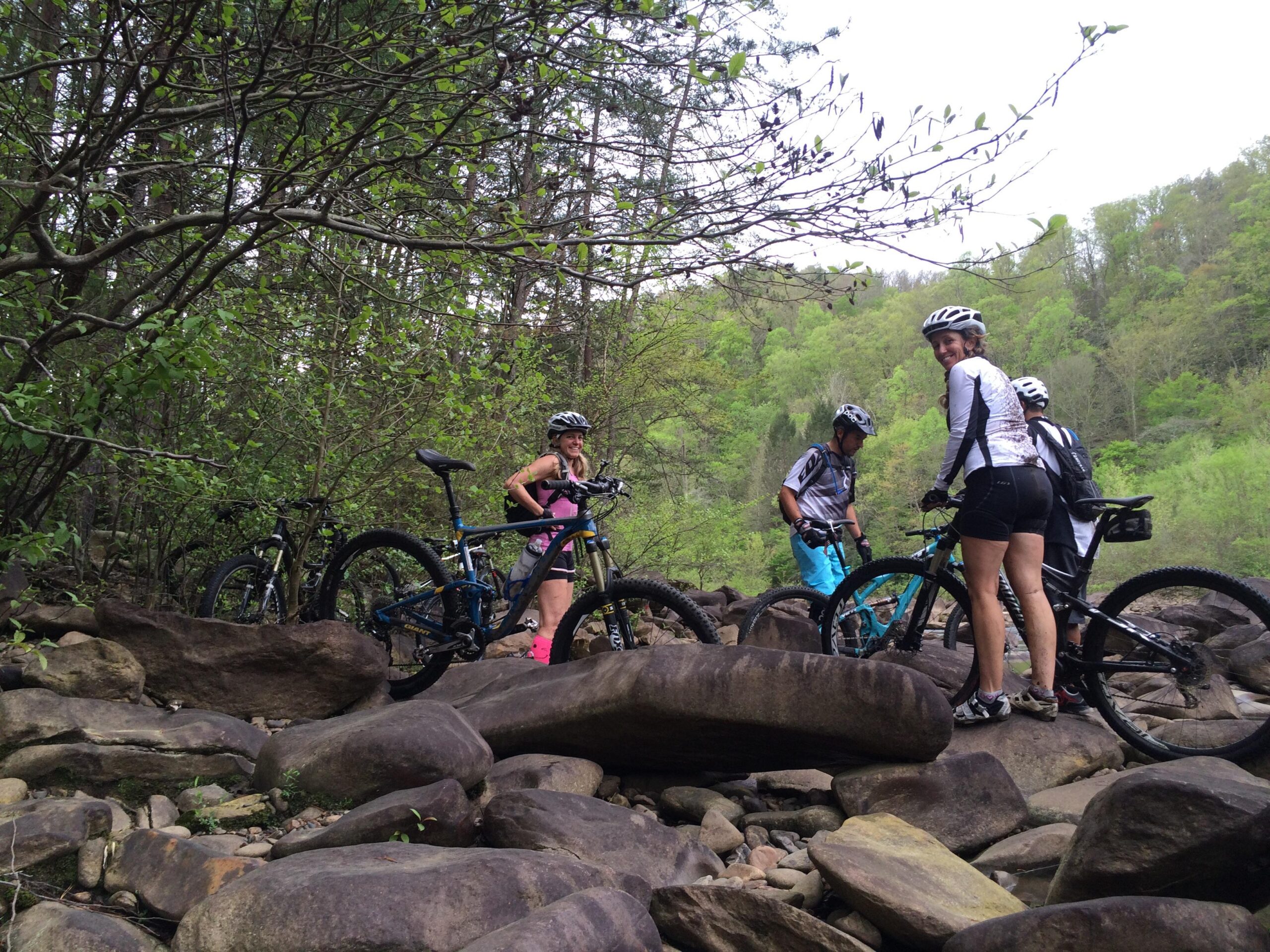 Group of mountain bikers navigating rocky terrain in a forested area, with bicycles positioned around them. The landscape features lush greenery in the background and boulders scattered on the ground. Tanasi Trail System mountain bike trail.