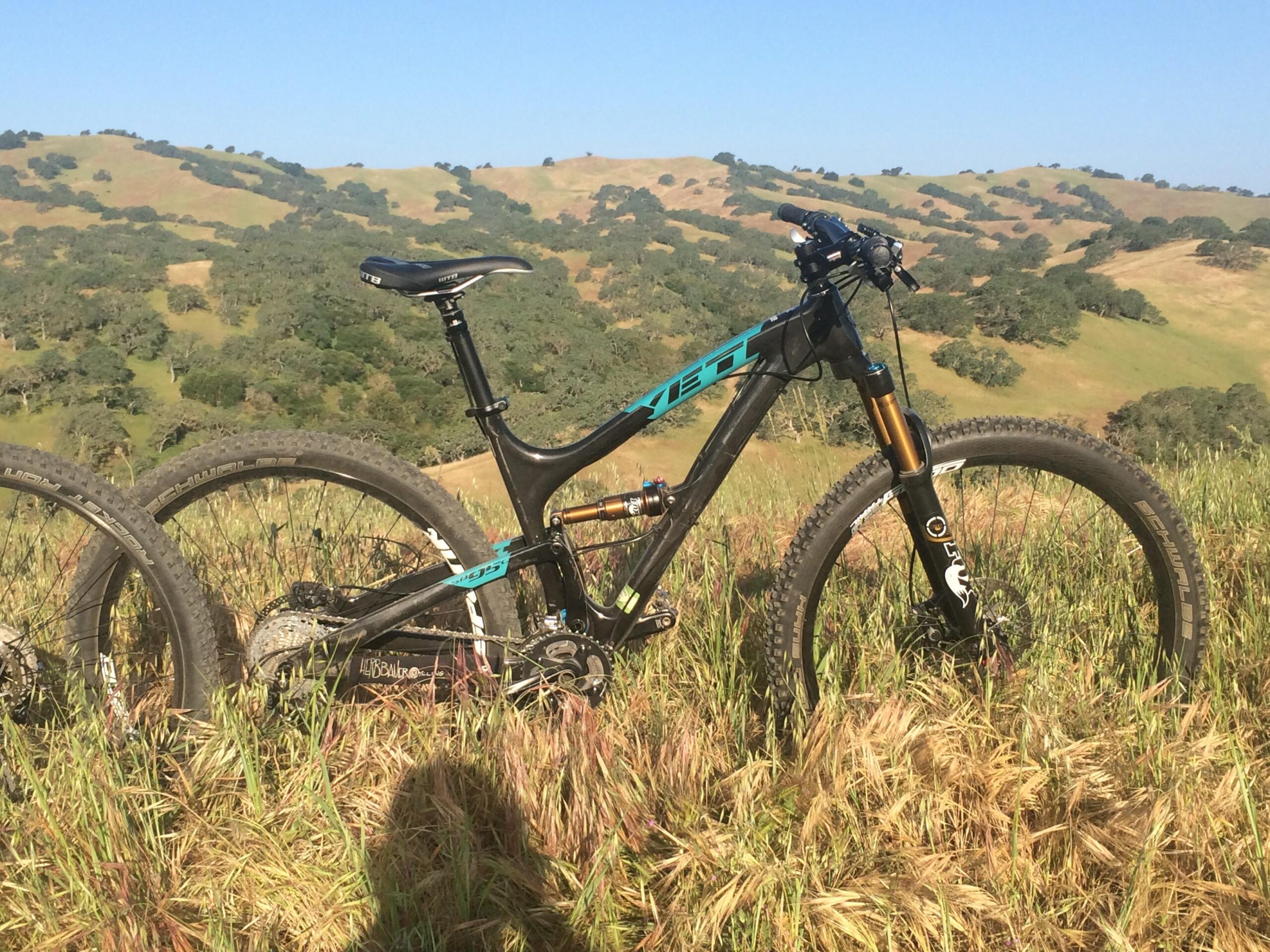 Yeti SB95: A mountain bike resting on a grassy hillside, with rolling green hills and trees in the background under a clear blue sky. The bike features a sleek black frame with turquoise accents and wide tires designed for off-road terrain.