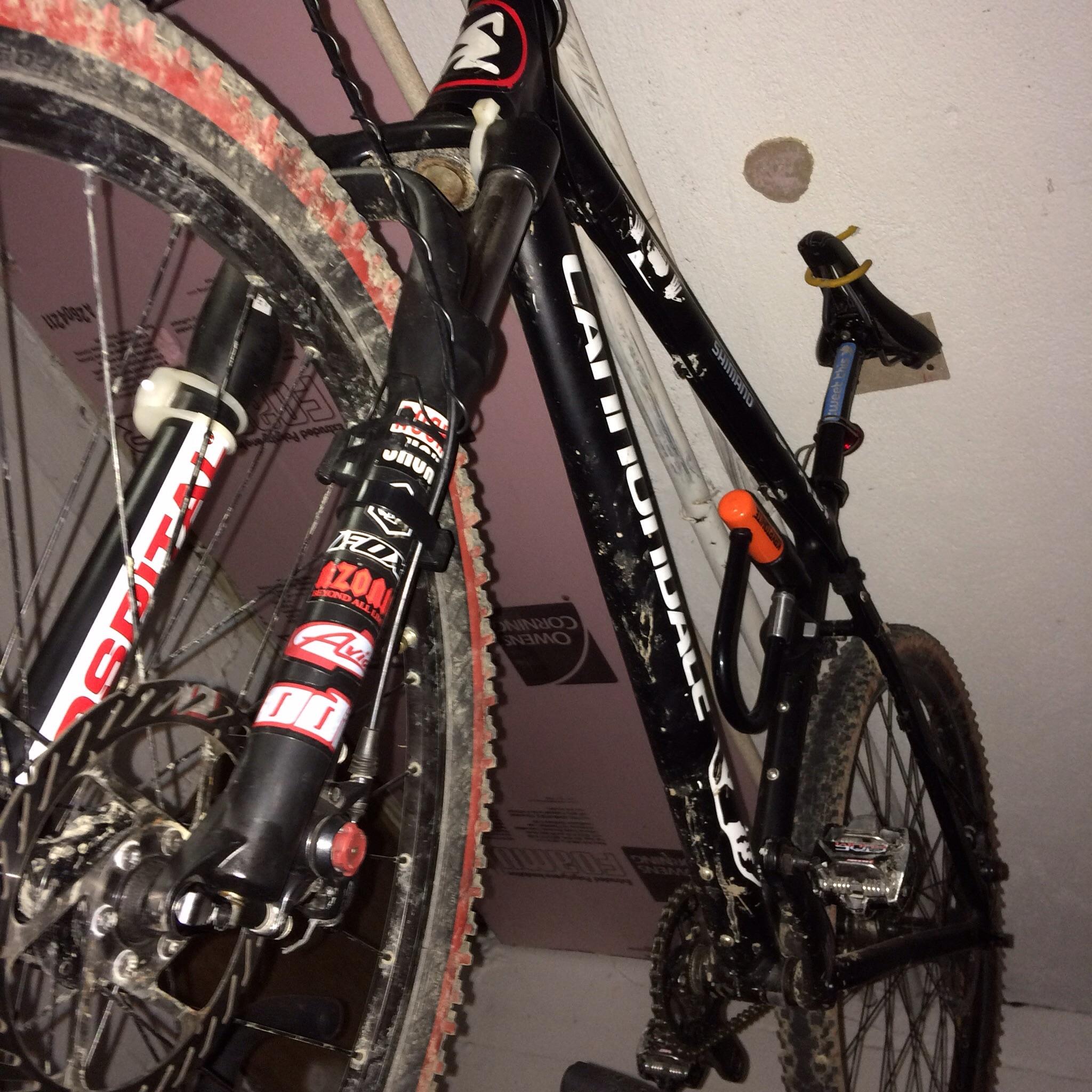 Cannondale F5: A black mountain bike with visible dirt and wear, mounted upside down against a light-colored wall. The bike features distinctive branding and stickers, with details of the frame, handlebars, and wheels visible.