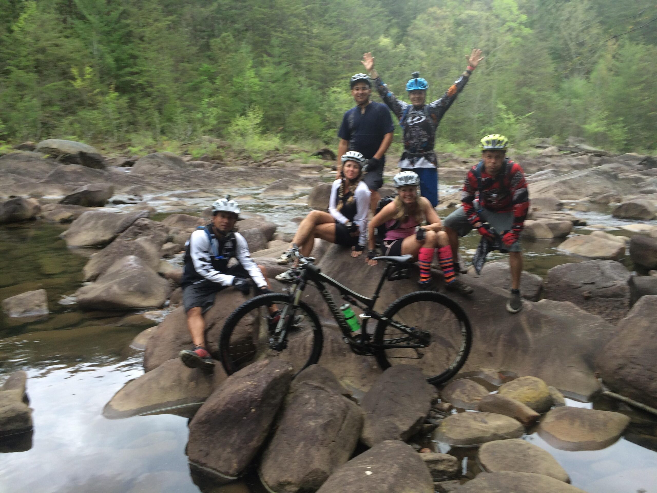 A group of six mountain bikers gathered on large rocks by a serene river, surrounded by lush greenery. Some are posing enthusiastically, while others are seated with their bikes next to them, enjoying a moment of camaraderie in nature. The scene captures a sense of adventure and outdoor fun. Tanasi Trail System mountain bike trail.